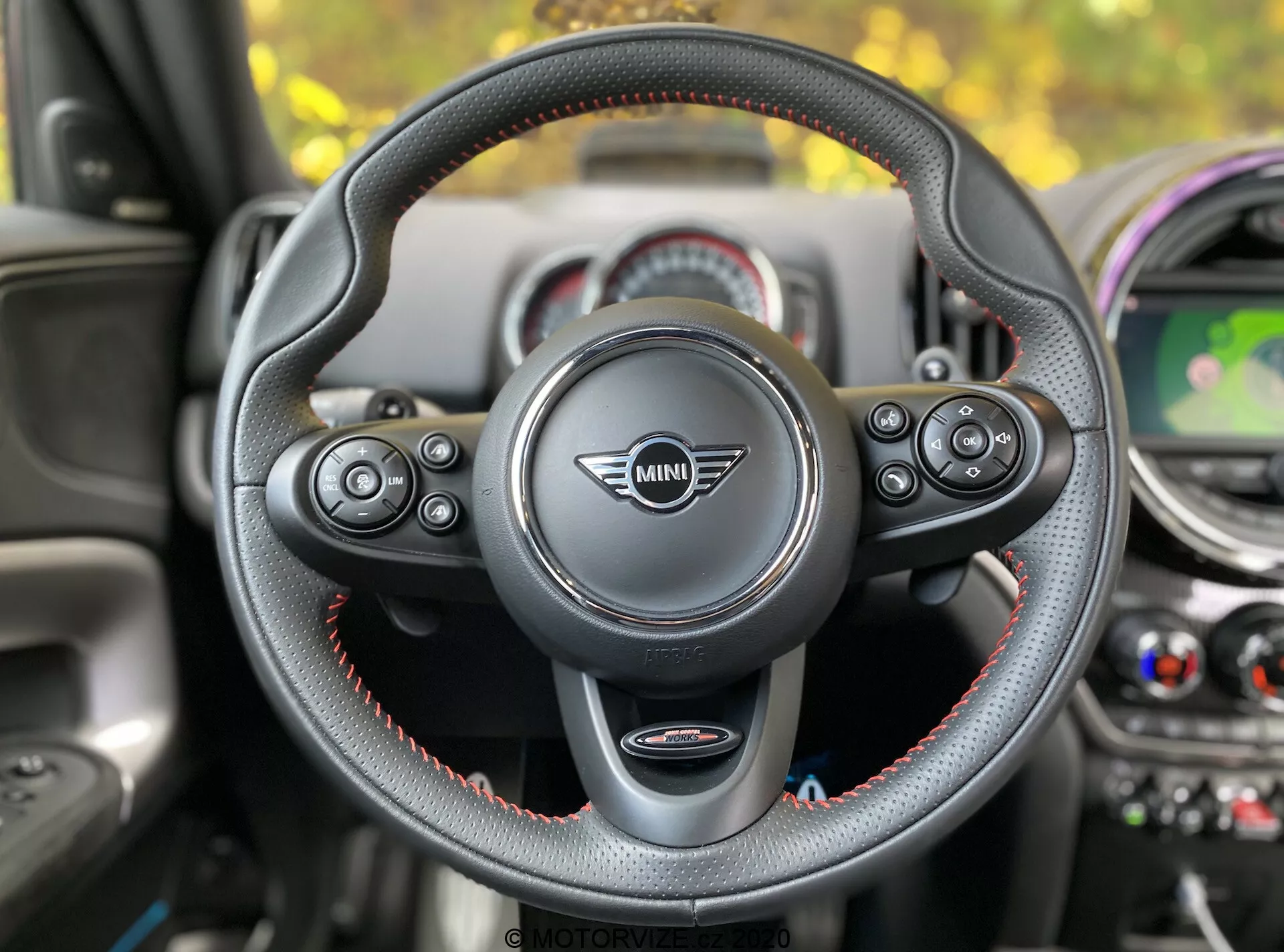 Vue intérieure d'une Mini Countryman John Cooper Works 2020 depuis le siège du conducteur, se concentrant sur le volant avec le logo Mini au centre. Le volant à trois branches, gainé de cuir avec des surpiqûres rouges, comporte des commandes multifonctions pour l'audio, le téléphone, le contrôle vocal et le régulateur de vitesse. Les palettes de changement de vitesse sont visibles, indiquant une transmission automatique avec sélection manuelle des rapports. En arrière-plan, on aperçoit le tableau de bord avec des éléments de design circulaires, un écran central affichant une carte, des bouches de climatisation et des accents colorés.