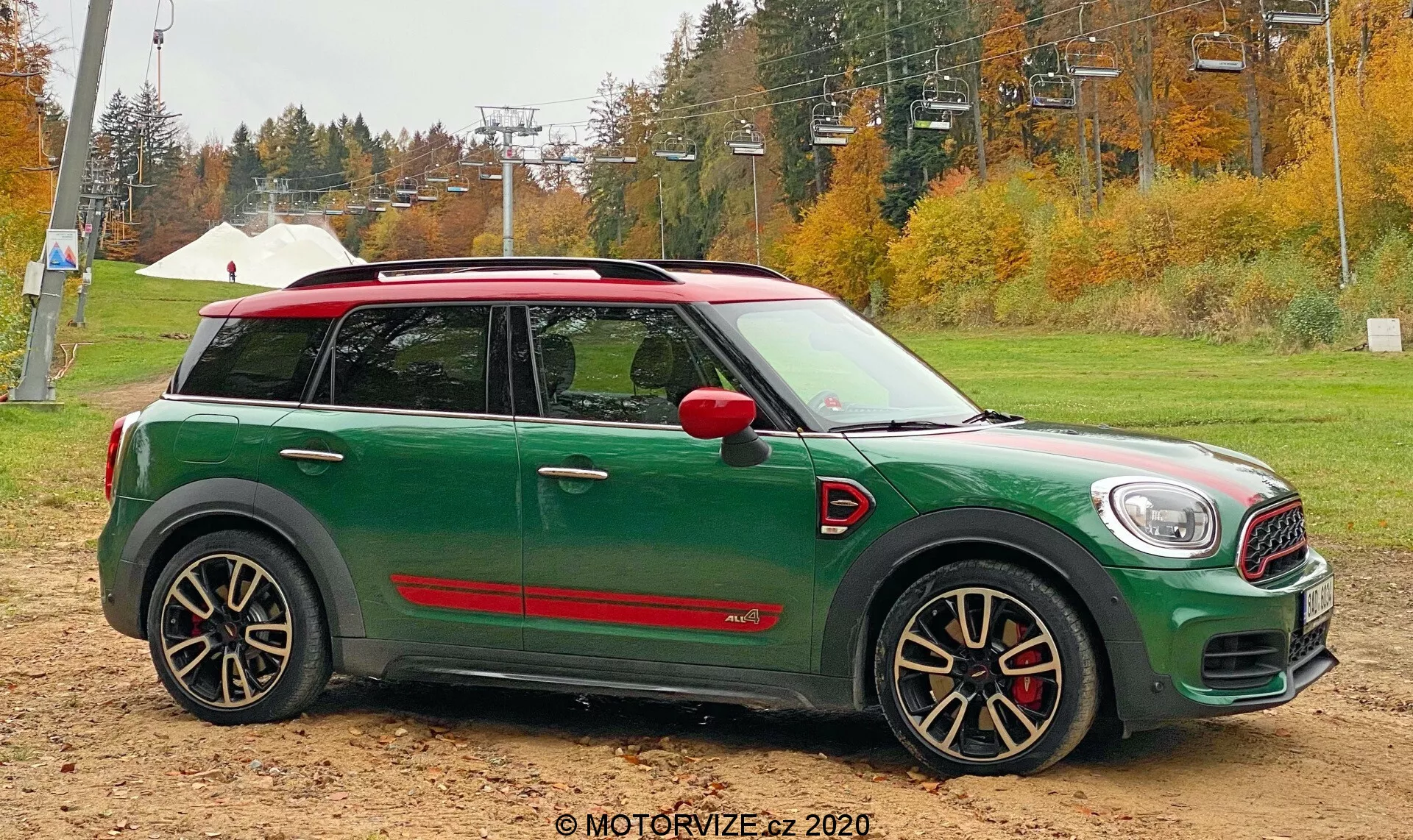 Profil latéral d'une Mini Cooper Countryman 2020 John Cooper Works verte, positionnée sur un sol sablonneux avec des arbres d'automne et une infrastructure de remontée mécanique en arrière-plan. La voiture présente un toit blanc, des coques de rétroviseurs rouges, des roues noires et argentées à plusieurs branches avec des étriers de frein rouges agressifs et des bas de caisse latéraux rouges sportifs.