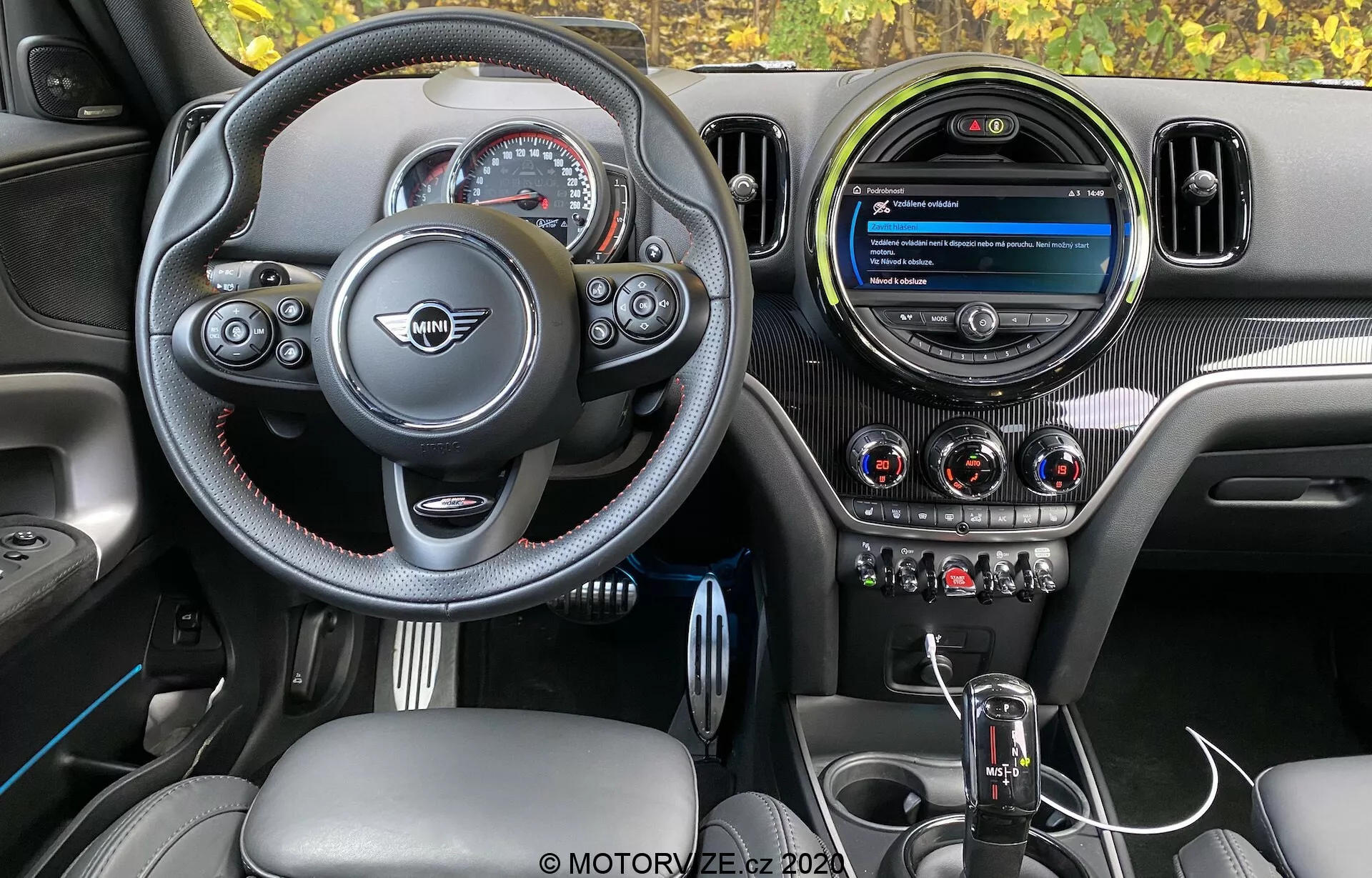 Vue intérieure du Mini Countryman John Cooper Works 2020, mettant en évidence le poste de conduite du conducteur avec un volant sportif au fond plat, des boutons multifonctions, des coutures contrastées et l'emblème central du fabricant. Le tableau de bord comprend un écran de divertissement circulaire moderne, trois bouches d'aération métalliques et des boutons de contrôle de la climatisation bien intégrés. La console centrale présente un levier de vitesses automatique avec un soufflet en cuir et un bouton de commande d'infodivertissement, le tout complété par des textures en fibre de carbone, du cuir et des accents métalliques pour un design premium orienté vers les performances.