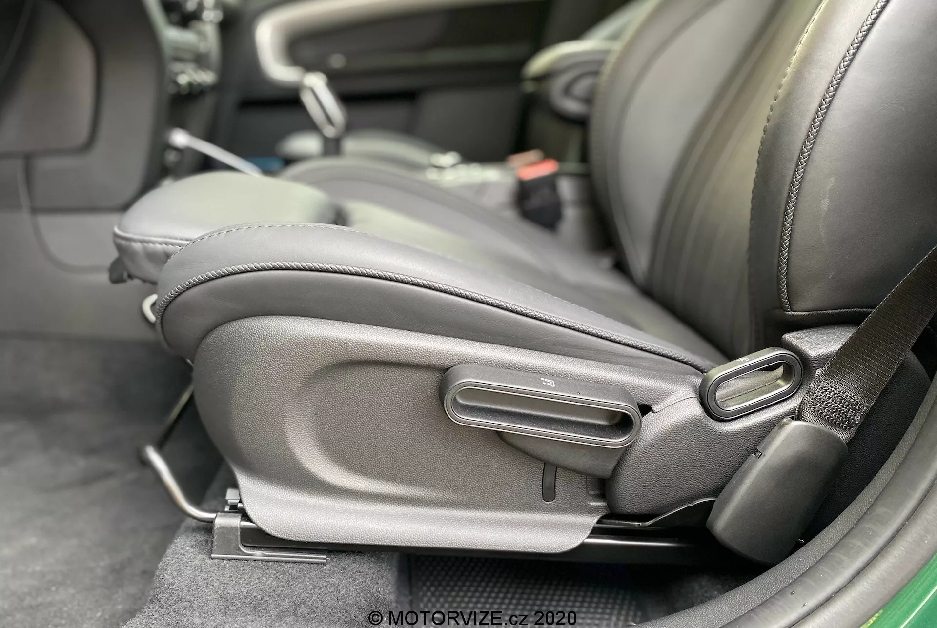 Vue rapprochée de l'intérieur de la Mini Countryman John Cooper Works 2020, mettant en évidence la partie inférieure latérale du siège du conducteur avec des mécanismes de réglage détaillés. Le siège est recouvert de cuir foncé ou de similicuir avec un renfort latéral proéminent, se concentrant sur le confort et la qualité. Les commandes de réglage manuelles, y compris une poignée en barre et un cadran rond, sont visibles, suggérant un design ergonomique et une ajustabilité utilisateur. Un réceptacle de ceinture de sécurité est également présent, indiquant une attention particulière à la sécurité des passagers.