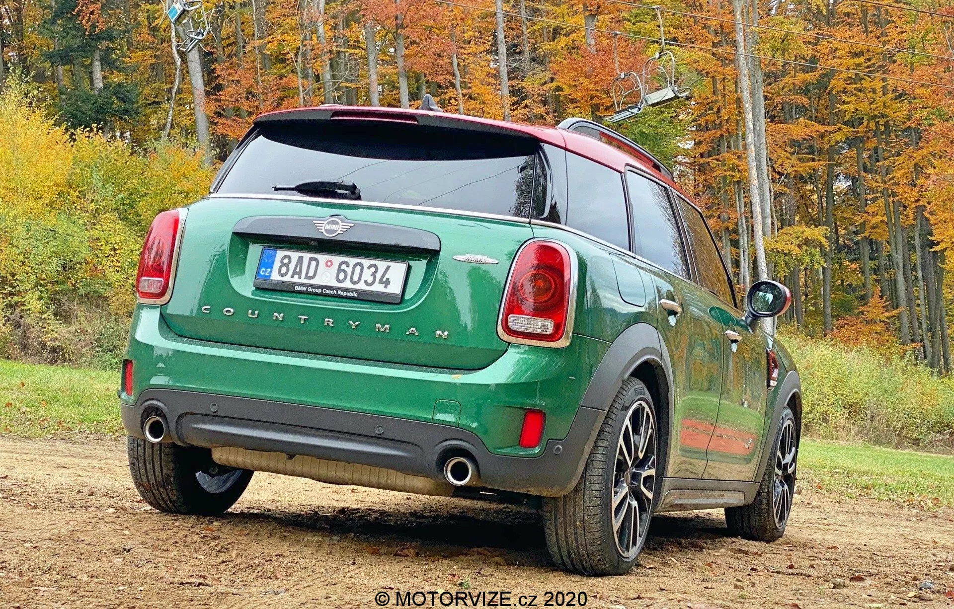 Image d'une Mini Countryman 2020 modèle John Cooper Works verte garée en plein air dans un décor automnal, prise sous un angle arrière trois quarts. Le côté arrière droit de la voiture est tourné vers la caméra et montre les feux arrière, le pare-brise arrière, le badge 'COUNTRYMAN', un système d'échappement double, des roues à cinq branches, un toit noir contrastant et des rétroviseurs latéraux. L'image inclut également une plaque d'immatriculation arrière, un essuie-glace arrière et une antenne pour la connectivité.