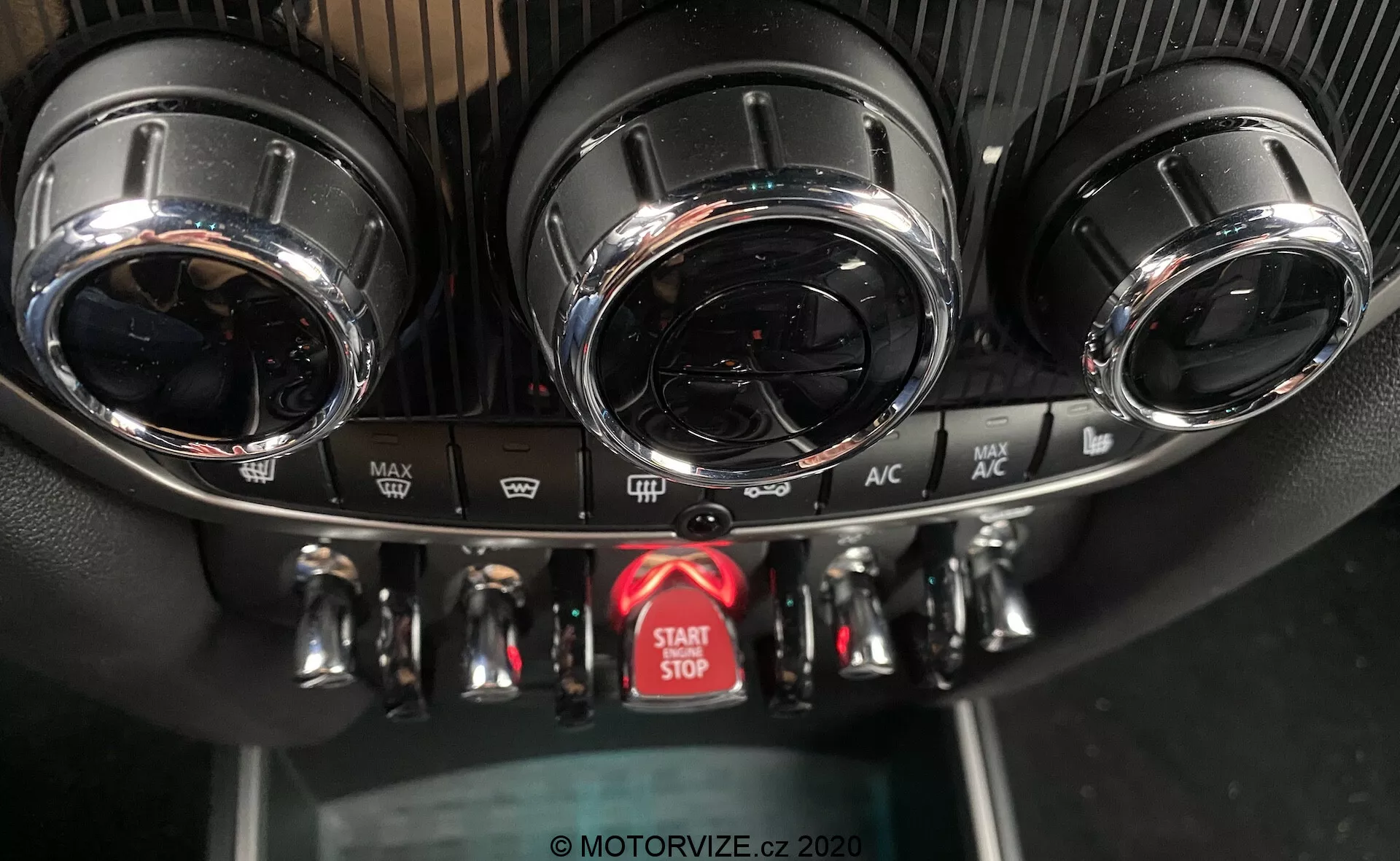 Cette image capture une vue en gros plan de la console centrale du tableau de bord d'une Mini Countryman John Cooper Works 2020, mettant en évidence le système de contrôle du climat et le bouton de démarrage/arrêt du moteur. La console présente deux aérations circulaires avec des finitions métalliques en haut, un panneau de contrôle du climat détaillé avec trois cadrans métalliques pour la température, la vitesse du ventilateur et la direction du flux d'air, et plusieurs boutons pour des fonctions telles que le dégivrage et la recirculation de l'air. Sous les commandes se trouve un bouton rouge proéminent pour le démarrage et l'arrêt du moteur. Les textures du panneau comprennent un design sportif semblable à la fibre de carbone autour des aérations et du plastique noir standard pour le panneau inférieur. L'angle de la photo, de face et légèrement en hauteur, met en évidence le design haut de gamme et les caractéristiques avancées.