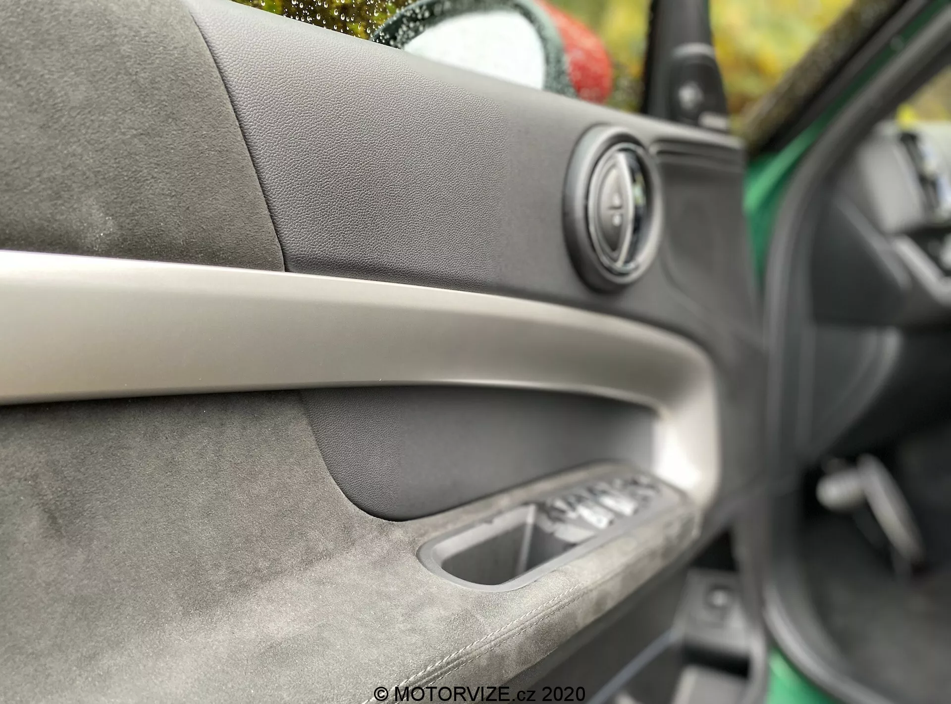 Photo de détail intérieur d'une porte de voiture Mini Countryman John Cooper Works 2020. Focalisée sur un haut-parleur audio premium avec anneau métallique, bande décorative diagonale, matériau doux au toucher avec coutures visibles, poignée de porte intérieure et commandes des vitres électriques. Profondeur de champ réduite mettant en avant l'artisanat et le confort intérieur.