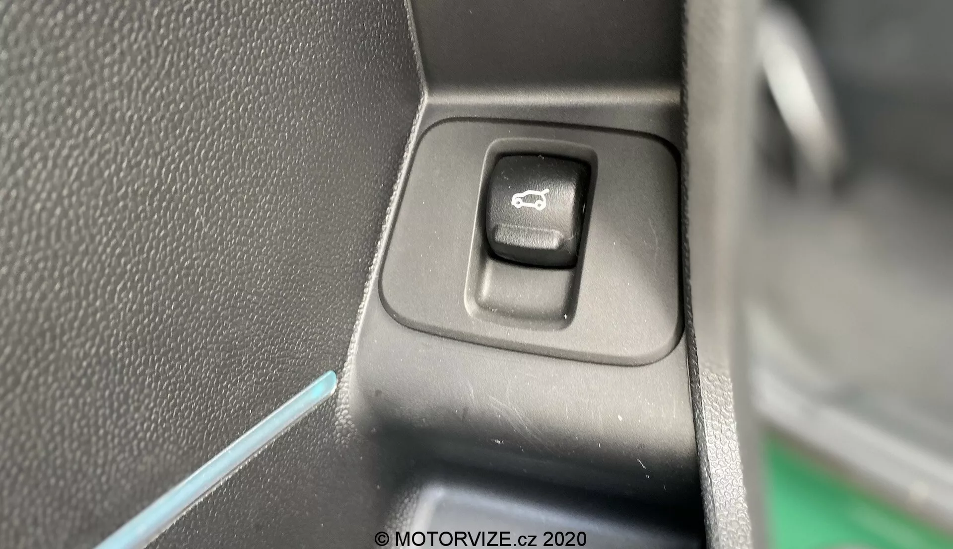 Vue rapprochée de l'interrupteur de commande pour les rétroviseurs latéraux à l'intérieur du Mini Countryman 2020 John Cooper Works. L'interrupteur rectangulaire aux coins arrondis, situé sur un panneau plat, est mis en évidence avec un symbole de miroir. Un côté de l'interrupteur est dans l'ombre, indiquant une source de lumière provenant du côté opposé. L'arrière-plan présente du plastique texturé noir et une fine ligne de lumière ambiante bleue, contribuant à l'esthétique intérieure du véhicule.