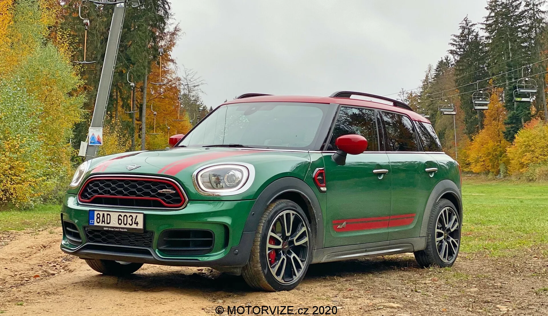 Image d'une Mini Countryman John Cooper Works 2020 verte dans une zone de gravier hors route avec un paysage forestier. Le crossover compact présente une vue avant trois quarts avec un toit et des rétroviseurs latéraux contrastants rouges, une grande calandre avant avec bordure chromée, des feux de jour à LED encadrant les phares principaux et des phares antibrouillard intégrés dans le pare-chocs inférieur. Les caractéristiques sportives notables comprennent une prise d'air sur le capot, des garnitures d'arches de roues noires, des jupes latérales, un spoiler arrière, de grandes roues en alliage à multiples rayons avec des pneus à profil bas et des étriers de frein rouges. Un toit panoramique et des rails de toit aérodynamiques sont également visibles.