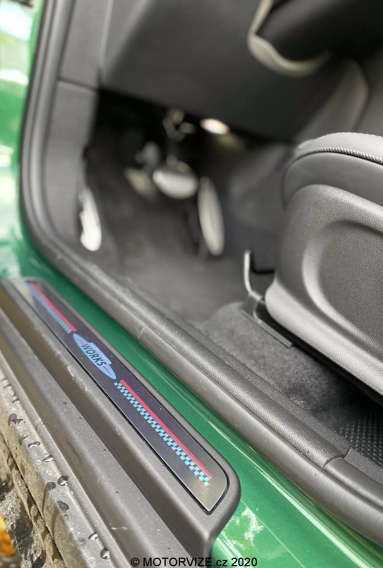 Vue rapprochée de l'aire du seuil de la porte de la Mini Countryman John Cooper Works 2020 sous un angle oblique focalisé sur le seuil du côté passager. L'image, capturée de l'extérieur du véhicule, met en évidence la partie inférieure de l'intérieur de la voiture, incluant la plaque de seuil avec des accents bleus et rouges, des tapis noirs, le montant latéral sportif du siège noir, une vue partielle des pédales et une zone de seuil bien entretenue. Les éléments extérieurs visibles incluent une jupe latérale verte et la carrosserie, rehaussant l'attrait esthétique. Le filigrane 'MOTORVIZE.cz 2020' suggère que la photo a été prise en 2020 pour une revue automobile.