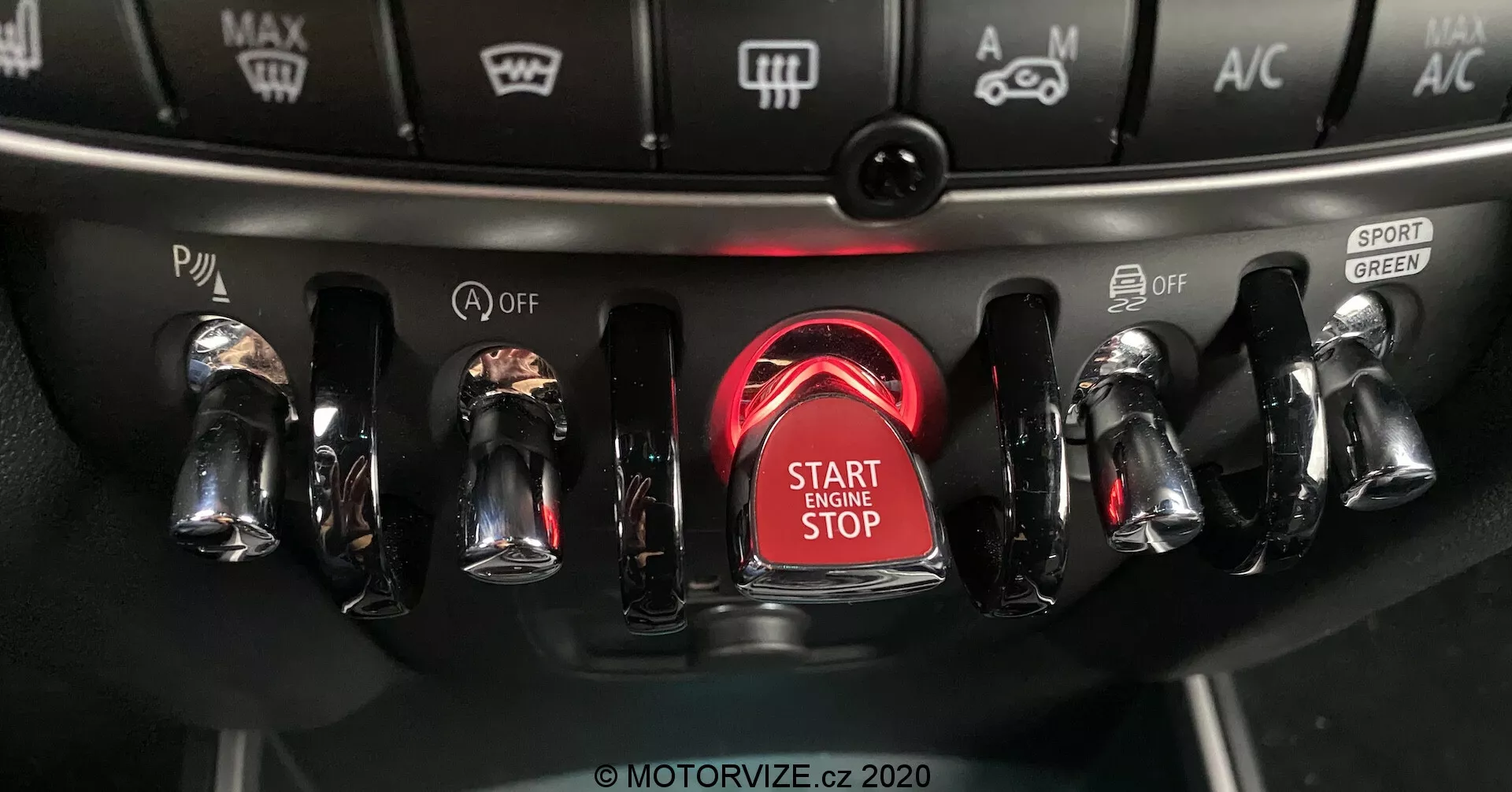 Vue en gros plan de la console centrale du Mini Countryman John Cooper Works 2020, mettant l'accent sur la zone d'allumage avec un bouton de démarrage/arrêt du moteur en évidence entouré d'un anneau rouge brillant, comportant des commutateurs métalliques au fini chromé de chaque côté pour divers contrôles du véhicule, y compris les modes de conduite et les réglages climatiques.