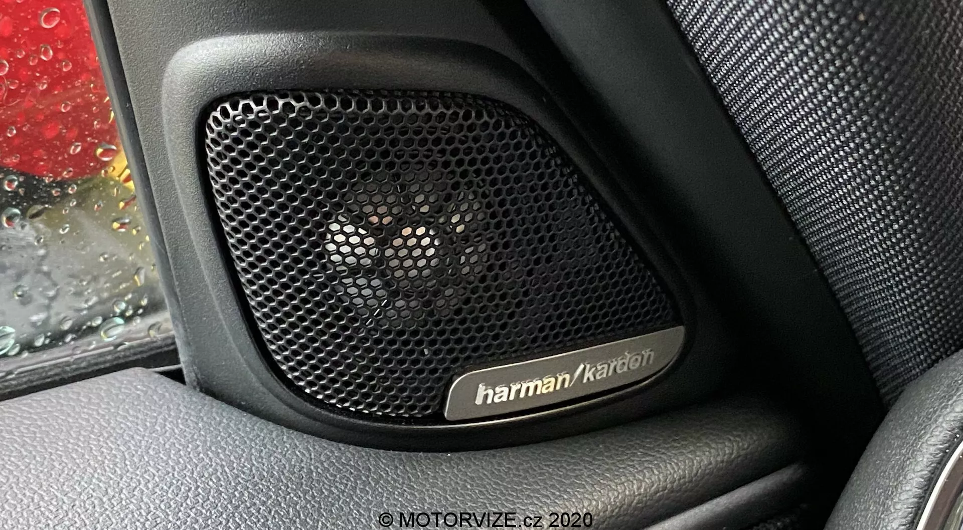 Vue rapprochée du système de haut-parleurs Harman Kardon à l'intérieur d'un panneau de porte de Mini Countryman 2020 John Cooper Works. La photographie se concentre sur la grille de haut-parleur perforée, conçue pour la transparence acoustique et la protection, contre un intérieur à texture cuir. Des gouttes de pluie sont visibles sur la vitre de la voiture, ajoutant un contexte à l'image. Le système audio premium signale des caractéristiques technologiques clés, mettant en valeur le luxe et la durabilité du design du véhicule.