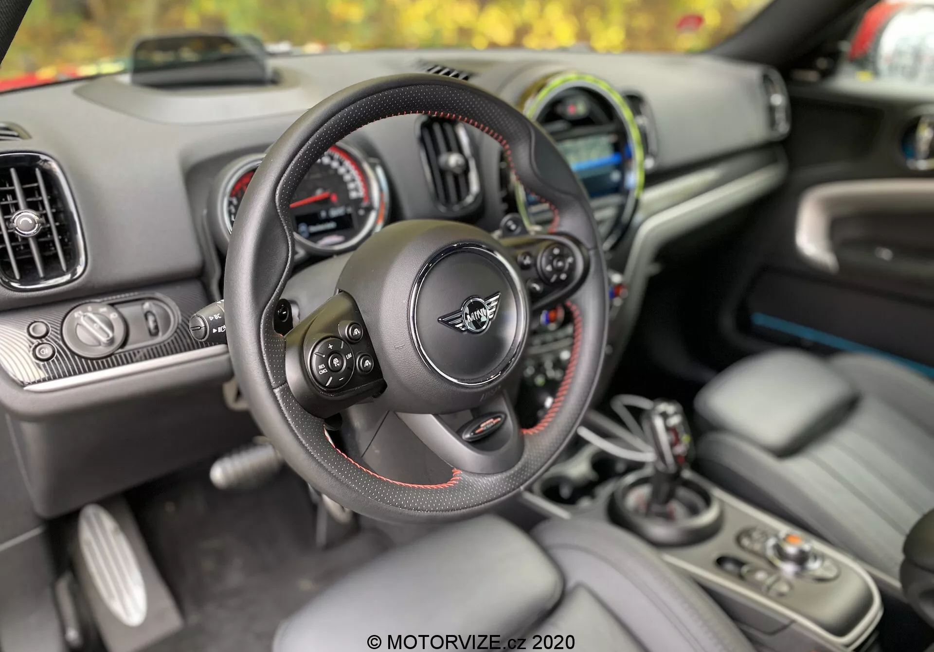 Vue intérieure de la Mini Countryman 2020 depuis la perspective du conducteur, mettant en évidence le volant multifonction avec des surpiqûres rouges, le tableau de bord avec des affichages analogiques et numériques, les bouches d'aération et l'écran d'infodivertissement. Sur la console centrale, on peut voir le levier de vitesses, les porte-gobelets, les boutons de commande et le porte-clés indiquant l'entrée sans clé et le démarrage par bouton-poussoir. L'avant-plan est centré sur le volant et le levier de vitesses, montrant l'ergonomie et le confort conçus pour le conducteur.