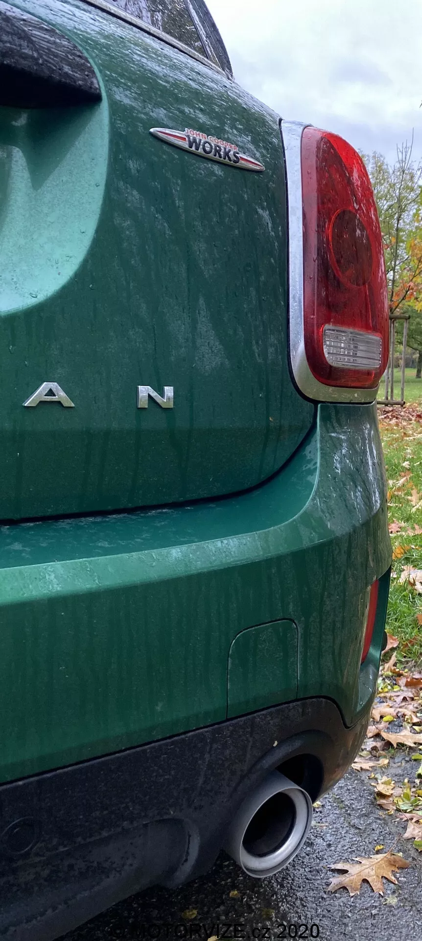 Vue rapprochée du coin arrière gauche d'une Mini Countryman John Cooper Works 2020 verte, mettant en évidence le feu arrière, les lettres chromées 'MINI', le badge 'WORKS', le pare-chocs arrière et l'échappement, indiquant des caractéristiques haute performance. L'image est prise par un jour d'automne nuageux, montrant des surfaces mouillées et des feuilles tombées.