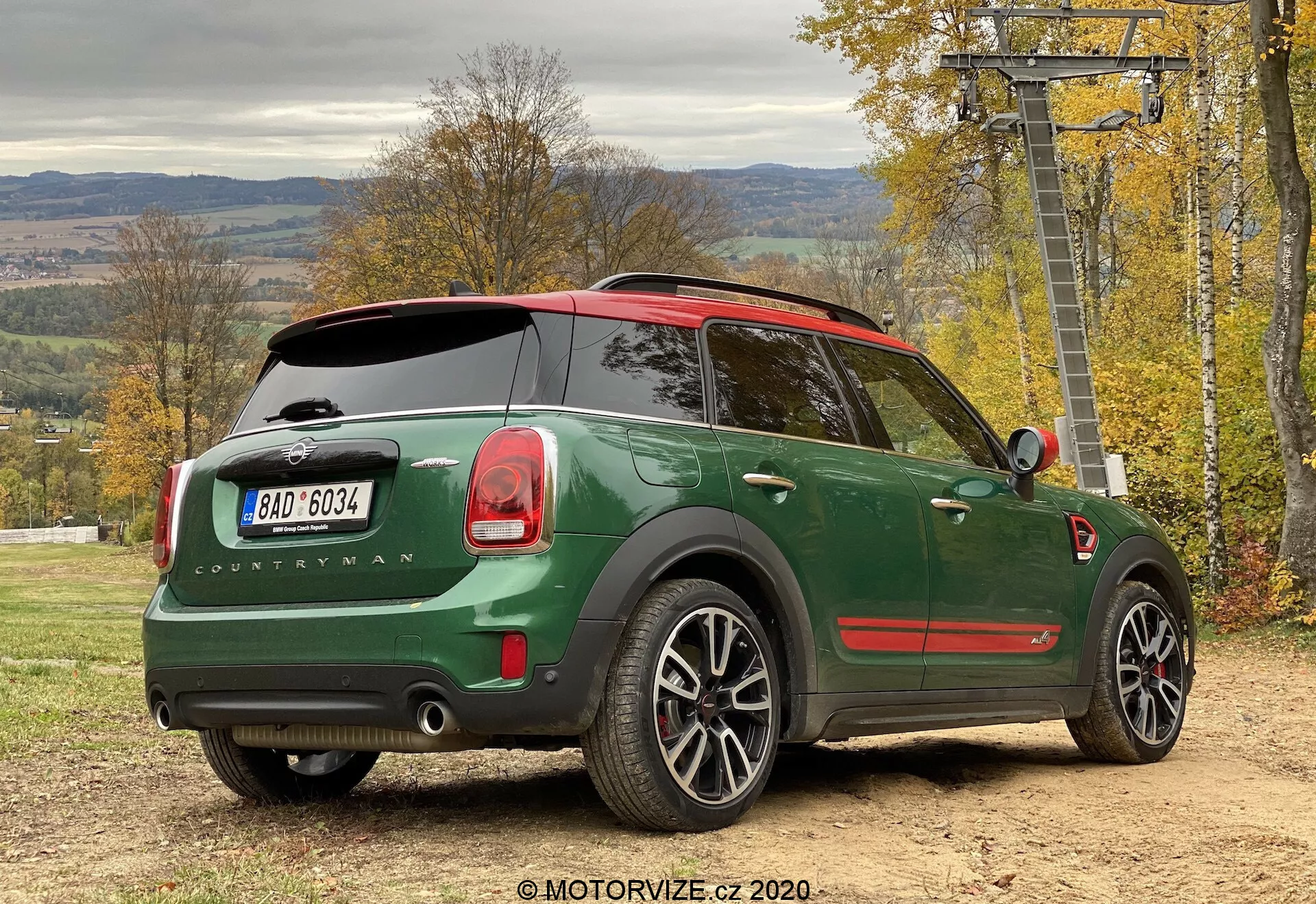 Vue arrière trois quarts d'une Mini Cooper Countryman John Cooper Works 2020 verte, garée en plein air, mettant en avant le design de la voiture depuis une perspective latérale arrière. Les détails notables incluent une carrosserie vert foncé avec un toit rouge, un badge 'Countryman', le logo Mini sur le hayon arrière, des feux arrière distinctifs, un système d'échappement double, des roues en alliage, un toit panoramique, un essuie-glace arrière, des fenêtres arrière teintées, des capteurs de stationnement et un rétroviseur latéral avec clignotant intégré. Le véhicule est sur une surface non pavée avec des feuillages d'automne et une structure de télésiège en arrière-plan, suggérant un style de vie actif et en plein air.