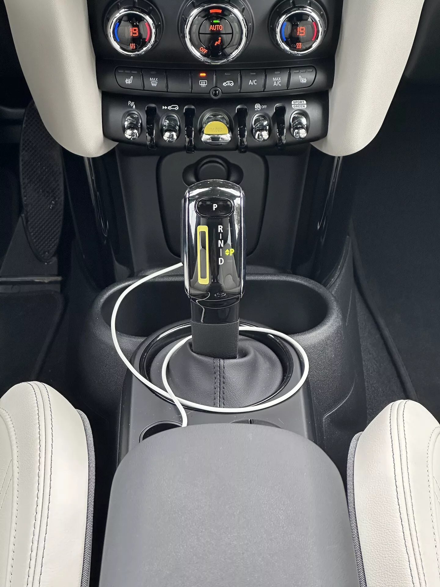 Vue rapprochée de la console centrale du Mini Electric 2021, mettant en évidence le sélecteur de vitesse en position 'P' avec une finition noire brillante et chrome et des indicateurs LED, entouré d'une surface noire mate avec un anneau argenté, des ports de connectivité USB, et zone de contrôle climatique avec des boutons métalliques et un affichage numérique de la température, le tout contre un design intérieur premium avec des couleurs de garniture contrastantes.