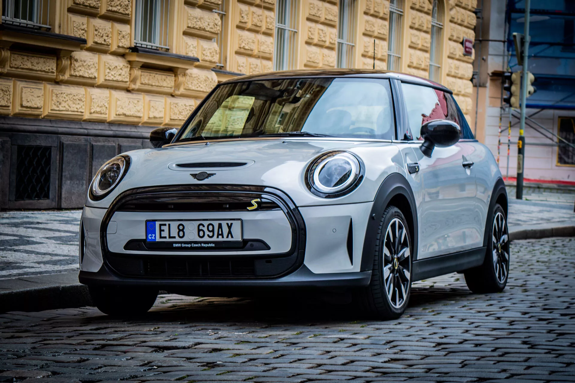 Mini Électrique 3 portes 2021 dans un cadre urbain montrant vue de côté avant gauche, phares LED circulaires, grille avant minimaliste avec emblème, roues en alliage foncé à design multibras, toit et coques de rétroviseurs contrastants, et esthétique sportive sur rue pavée