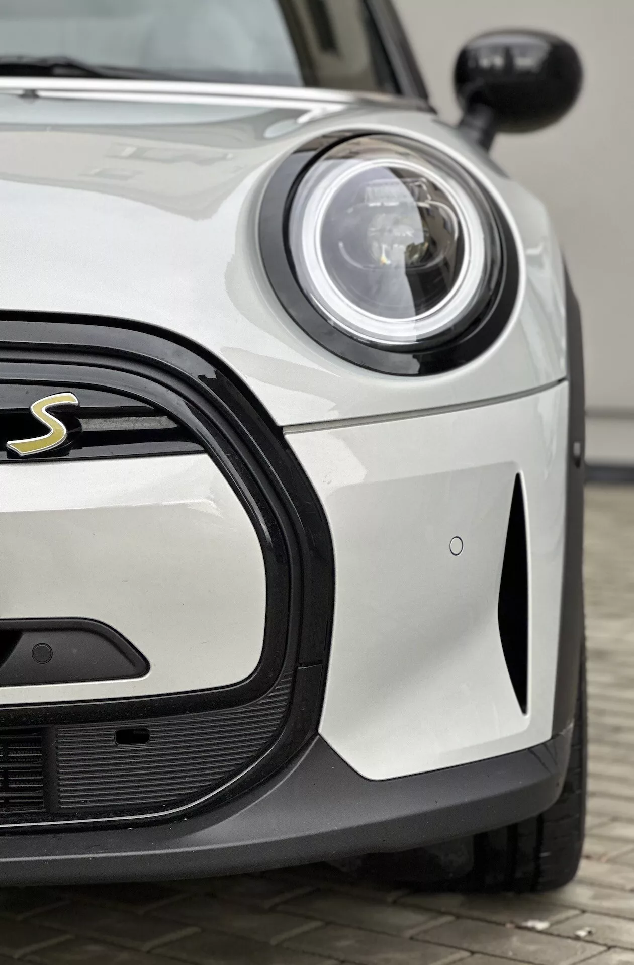 Gros plan sur le Mini Electric 3-Portes 2021 montrant le côté avant du conducteur, avec un phare circulaire LED ou Xénon à côté d’une grille élégante à motifs en nid d’abeille avec une vue partielle de l’emblème sportif, contre une carrosserie blanche solide