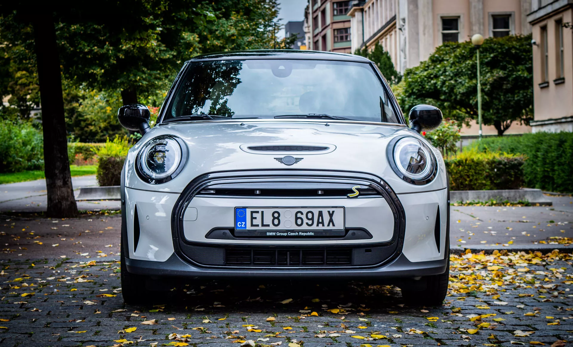 Vue de face d'une Mini Cooper 2021 Electric 3-portes sur une rue pavée avec des feuilles d'automne, présentant la technologie des phares LED, des capteurs d'aide au stationnement, un pare-chocs style sportif et le détail de l'emblème.