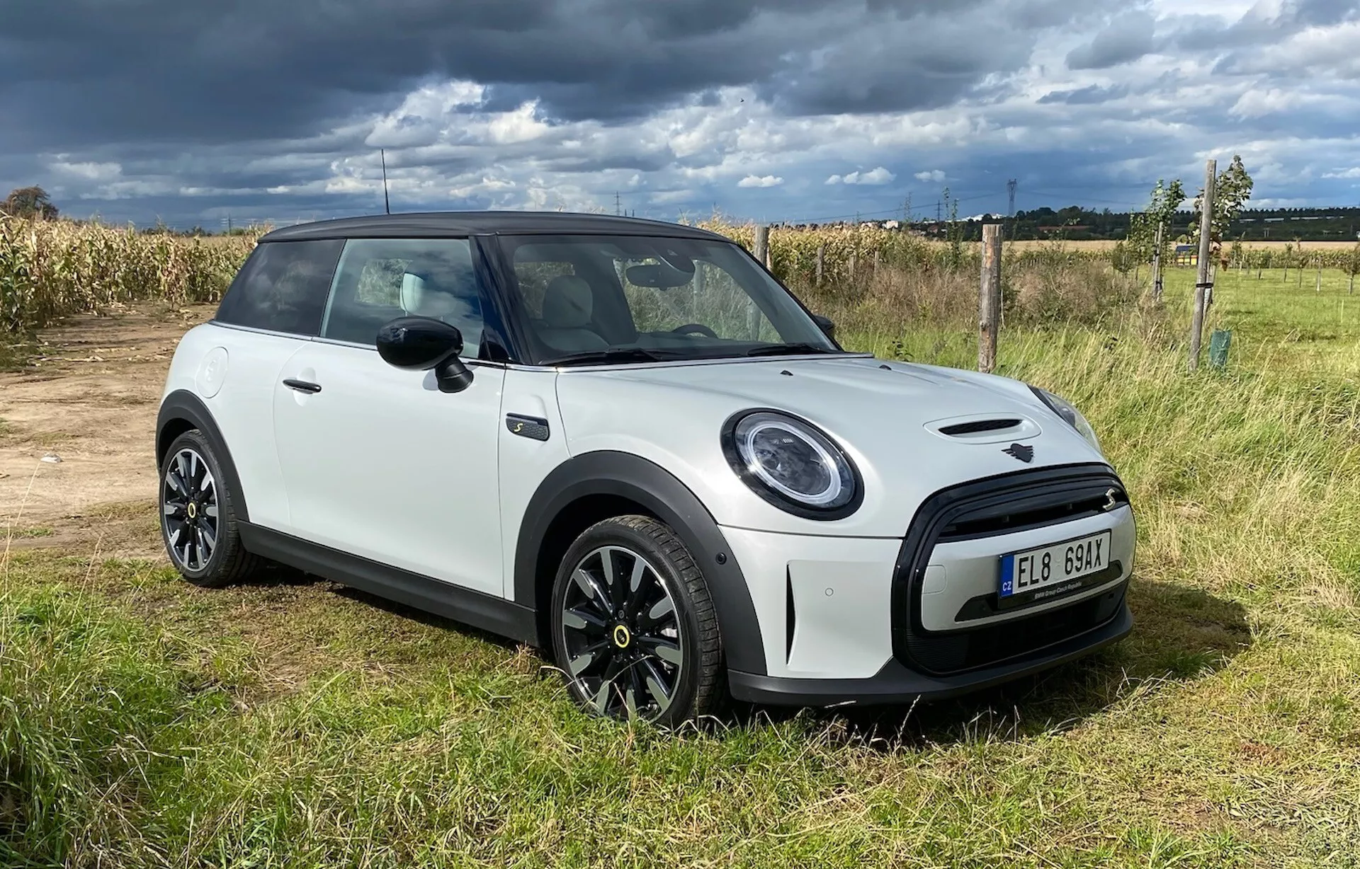 Modèle Mini Cooper Électrique 3 portes de 2021 en blanc, vue de face inclinée sur l'herbe avec ciel nuageux, présentant une garniture noire personnalisée, des phares arrondis, une calandre hexagonale et des jantes en alliage à plusieurs branches