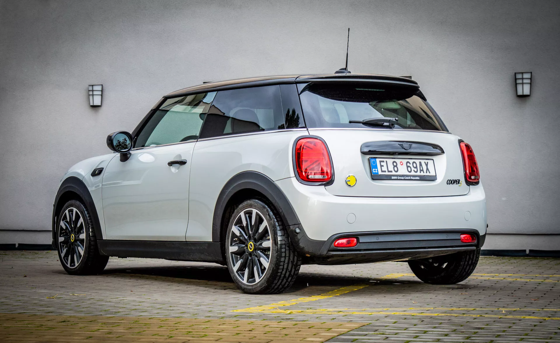 Hatchback Mini Électrique 3 Portes 2021 blanc vu de l'arrière trois quarts soulignant les feux arrière circulaires, l'insigne de la marque, et le pare-choc arrière avec garniture noire, équipé d'un essuie-glace arrière et d'un aileron de toit, profil latéral présentant des contours aérodynamiques, roues noires à plusieurs branches, poignées de porte chromées, et miroirs avec clignotants intégrés sur fond indistinct