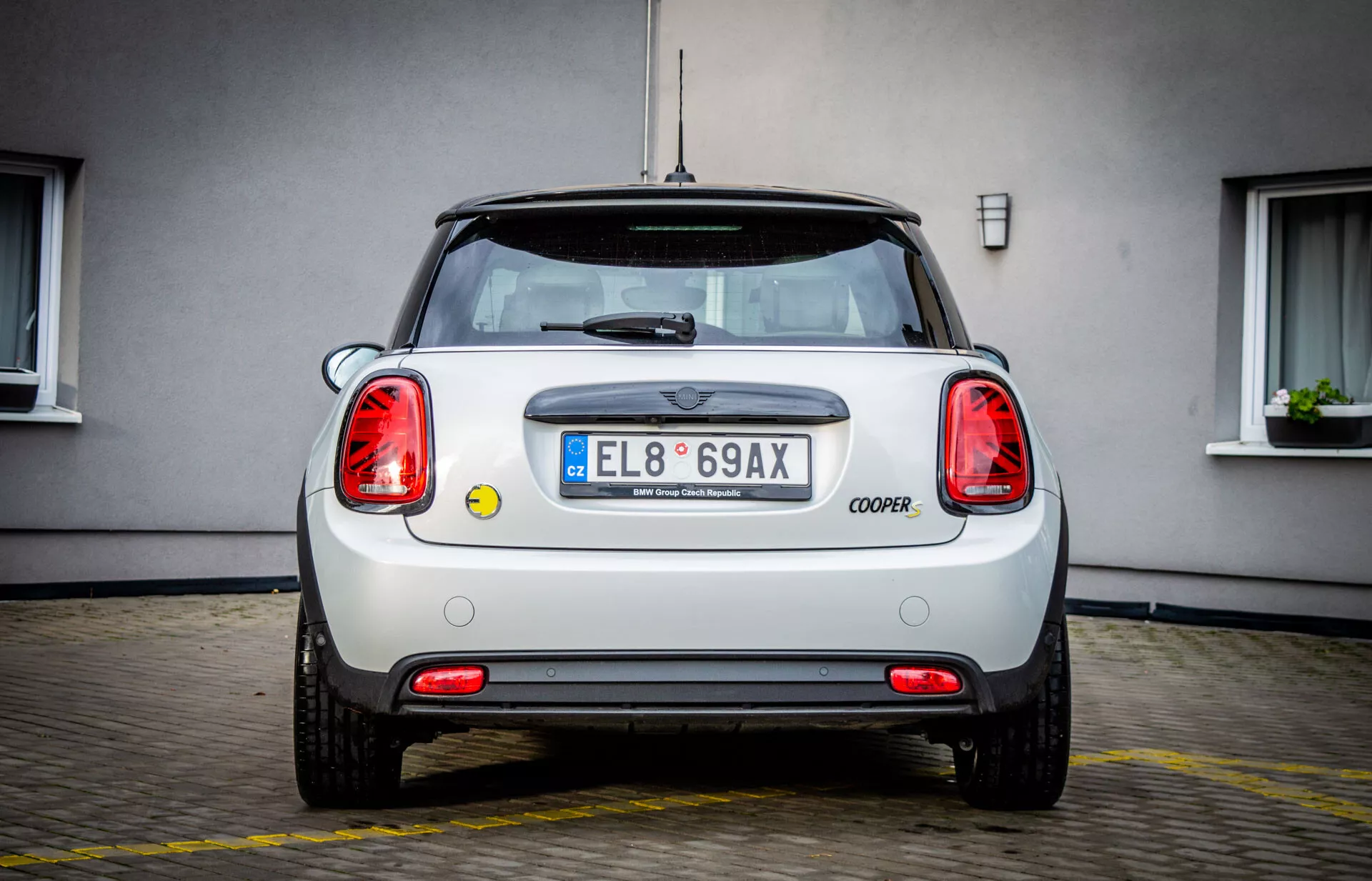 Vue arrière d'une Mini Electric 3-Door 2021 garée sur du béton, avec emblème, inscription COOPER, feux arrière circulaires, plaque d'immatriculation de l'UE avec 'E', pare-chocs impeccable avec réflecteurs, capteurs de stationnement, sortie d'échappement, schéma de couleur bicolore, antenne radio et lunette arrière avec essuie-glace
