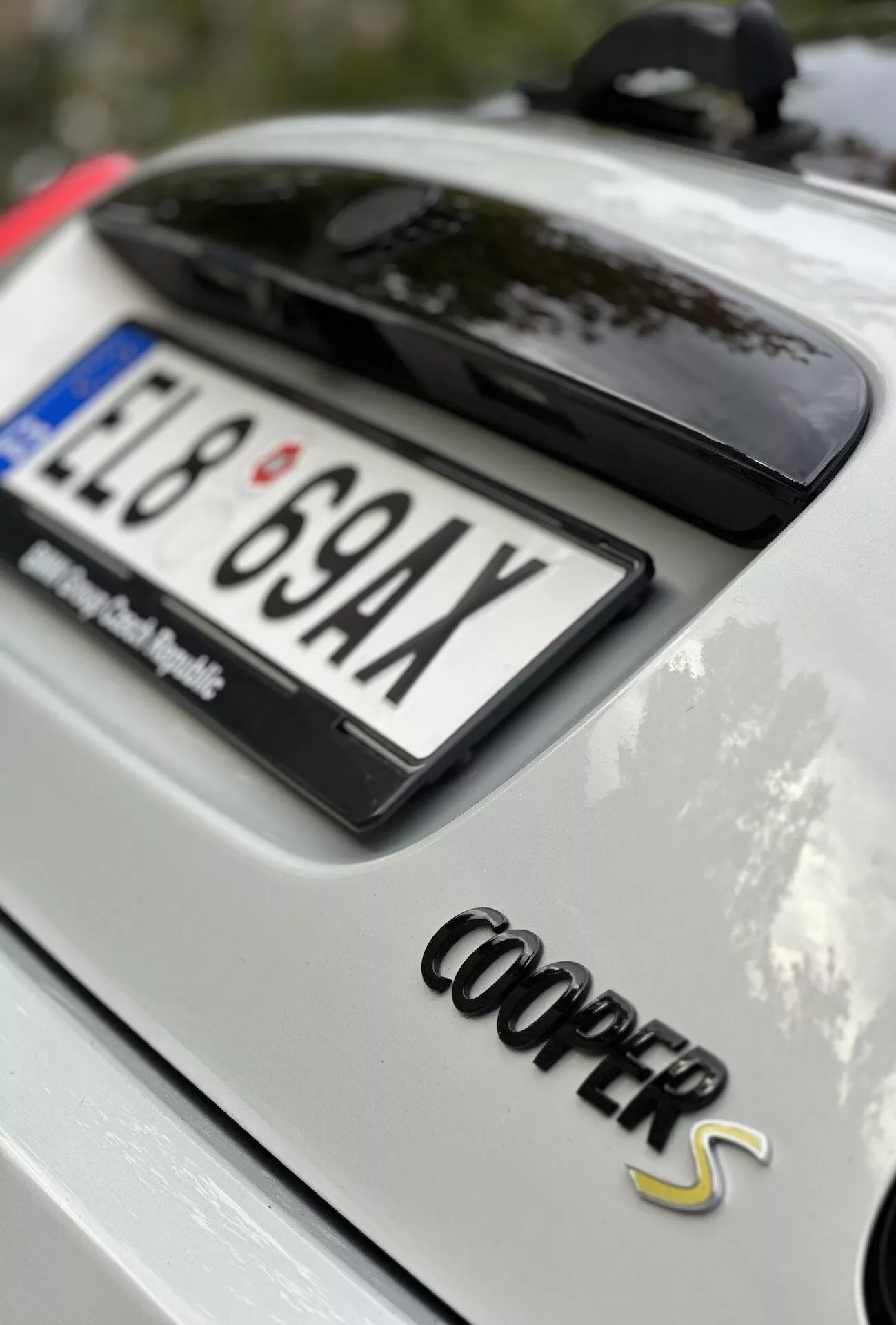 Vue arrière d'une Mini Cooper S Électrique 2021 blanche avec badge 'Cooper S' en chrome, plaque d'immatriculation européenne partielle et verre arrière teinté noir brillant ou toit ouvrant, montrant la zone du coffre de la voiture, antenne aileron de requin indiquant la technologie GPS, cadre extérieur