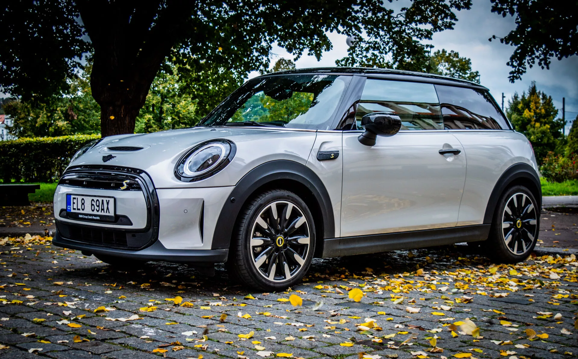 Mini Électrique 3 portes de 2021 en schéma de couleurs bicolore argent et noir, capturée sous un angle latéral bas dynamique mettant en évidence son design sportif, ses phares avant circulaires à LED, et ses jantes en alliage noir avec détails contrastés, stationnée dans une rue urbaine.