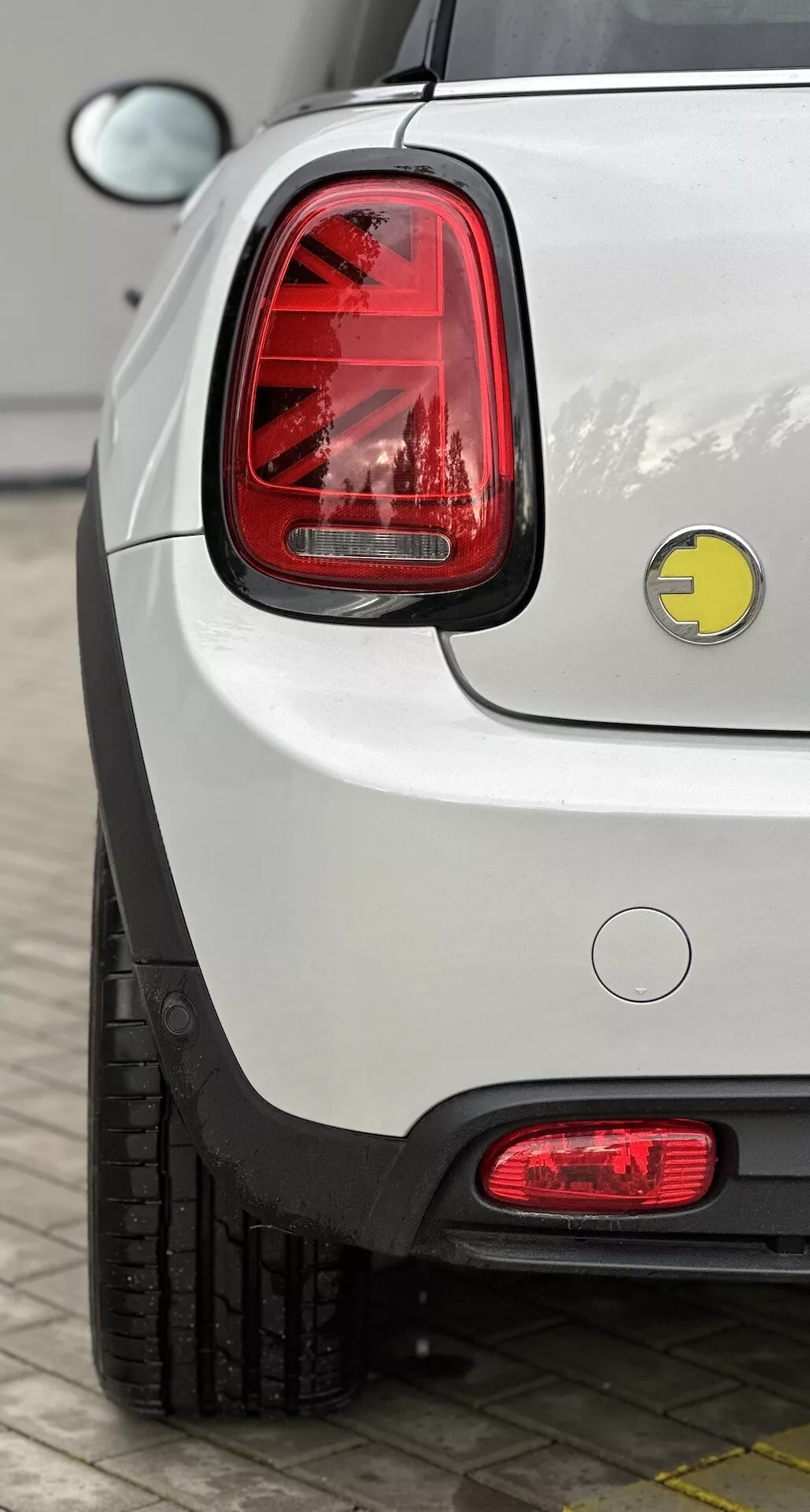 Vue rapprochée du coin arrière droit d'une Mini Electric 2021 blanche, mettant en évidence sa finition brillante, ses feux arrière LED verticaux, son badge de marque, son bouchon de carburant conventionnel, son pare-chocs arrière avec réflecteur, ses capteurs de stationnement, et la section visible du pneu, garée en extérieur.