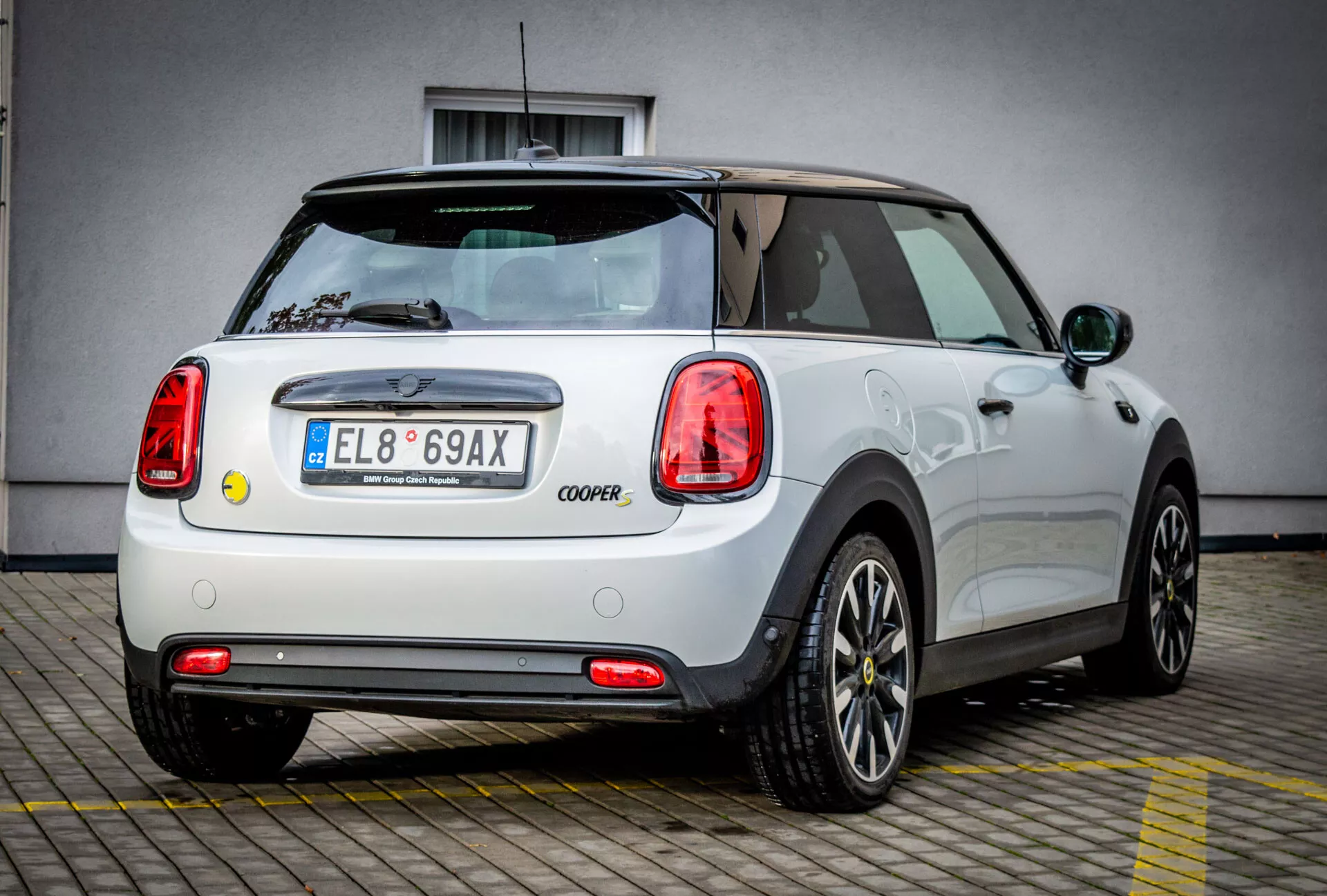 Vue arrière et de côté d'une Mini Electric 3-Door Cooper blanche de 2021, mettant en avant son design de roue distinctif et ses célèbres feux arrière cerclés de chrome, garée devant un bâtiment commercial