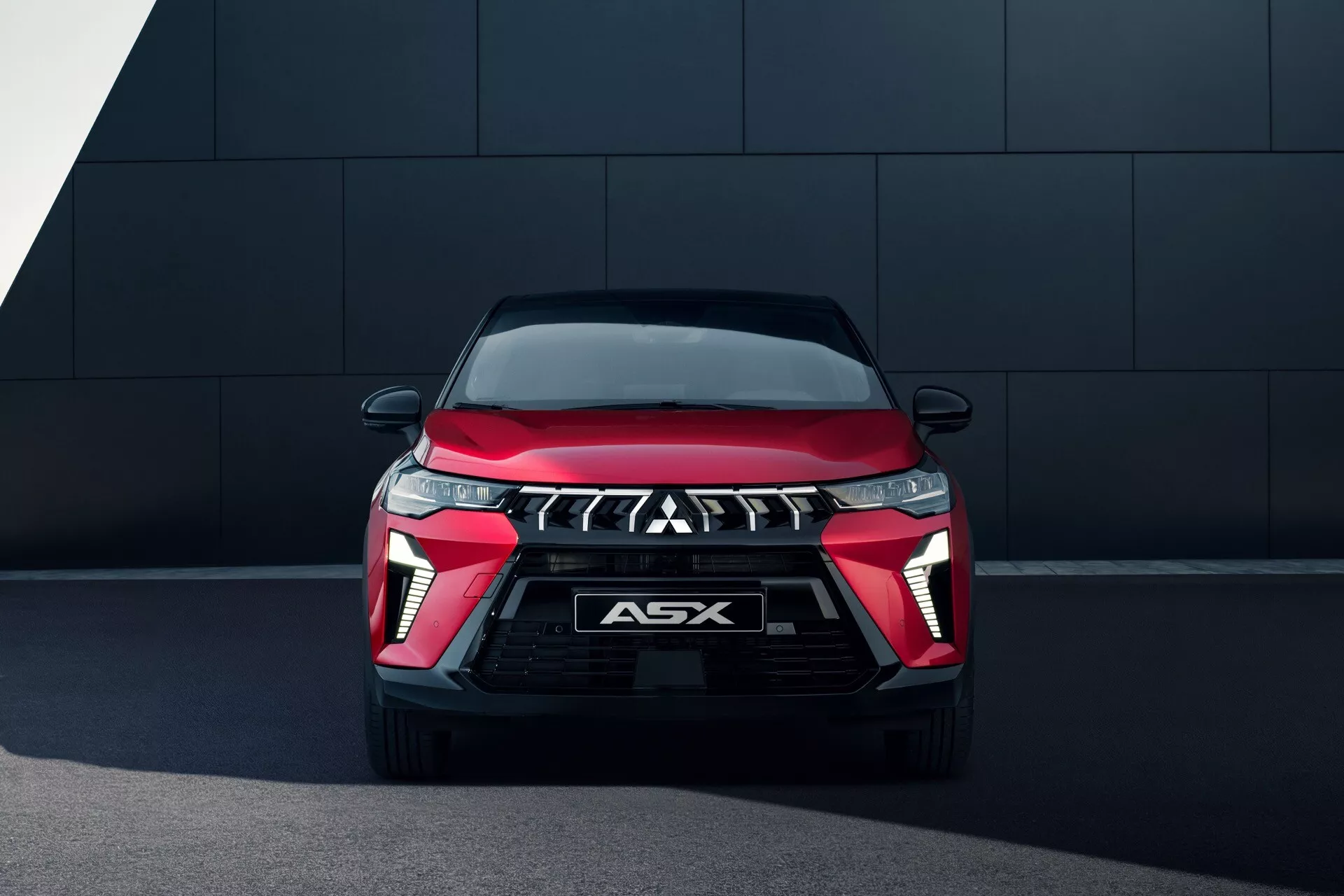 Vue frontale d'un Mitsubishi ASX 2024 (restylage de la 2ème génération), présentant son design moderne de SUV rouge et noir. La voiture est positionnée avec un léger angle vers le bas, mettant en valeur sa calandre distinctive à motif de drapeau à damier avec un emblème de marque proéminent. Encadrée par des phares LED élégants avec feux de jour intégrés et des logements de feux antibrouillard triangulaires, le véhicule affiche une esthétique high-tech et sportive. Le capot présente des lignes de caractère aérodynamiques et le pare-chocs avant noir comprend ce qui semble être des prises d'air. L'arrière-plan est un mur sombre et simple, soulignant les caractéristiques de conception dynamique de la voiture.