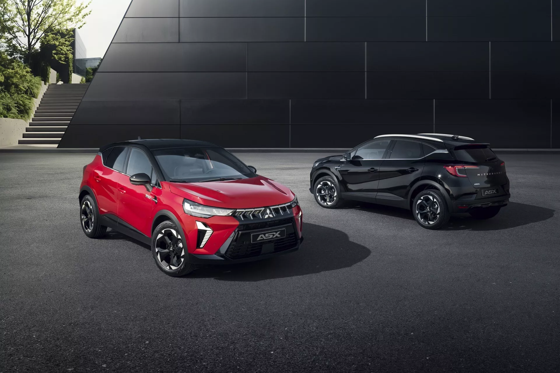L'image montre deux SUV dans un cadre extérieur avec une architecture moderne et minimaliste en arrière-plan. Au premier plan, on voit un Mitsubishi ASX 2024 rouge vif, positionné en diagonale par rapport à la caméra, affichant une vue de trois quarts avant qui met en valeur la calandre avant, les phares à LED, le pare-chocs avant, le capot et le profil latéral du conducteur incluant les roues, les portes et les rétroviseurs latéraux avec clignotants intégrés. En arrière-plan, un Mitsubishi ASX noir est visible sous un angle de trois quarts arrière, révélant les feux arrière, le hayon, le pare-chocs arrière, le profil latéral passager et la roue arrière avec des vitres teintées à l'arrière. L'éclairage professionnel accentue les lignes épurées, les profils aérodynamiques et les éléments de design moderne des voitures. Les technologies visibles incluent les feux diurnes à LED, les jantes en alliage, les capteurs de stationnement sur le pare-chocs avant du SUV rouge et des fonctionnalités avancées possibles comme le freinage d'urgence automatique ou le régulateur de vitesse adaptatif.