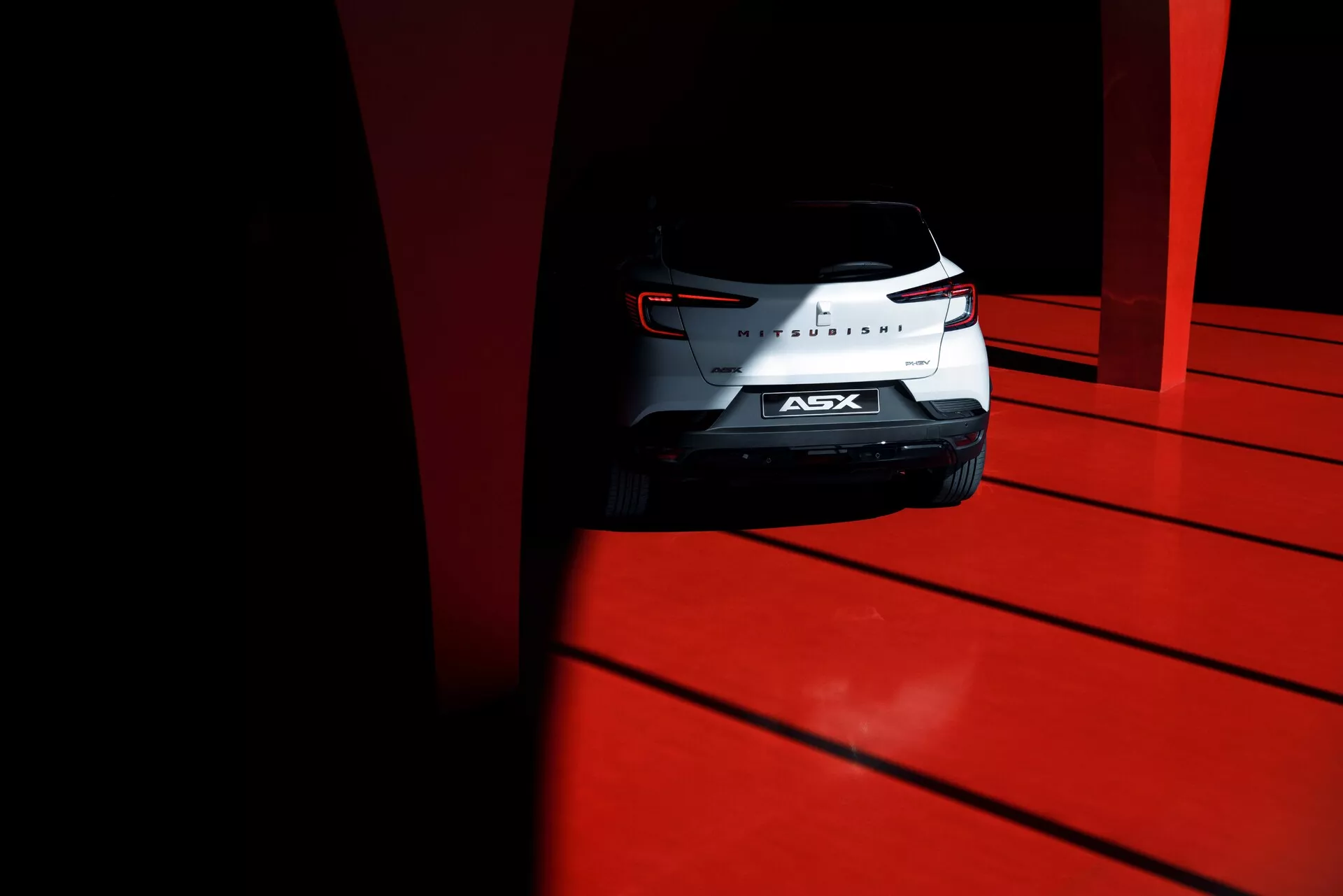 Prise en contre-plongée de l'arrière du SUV compact Mitsubishi ASX 2023, avec des feux arrière sculptés, un badge chromé et un pare-brise arrière, capturée en plein jour à haut contraste sur un sol rouge vif.