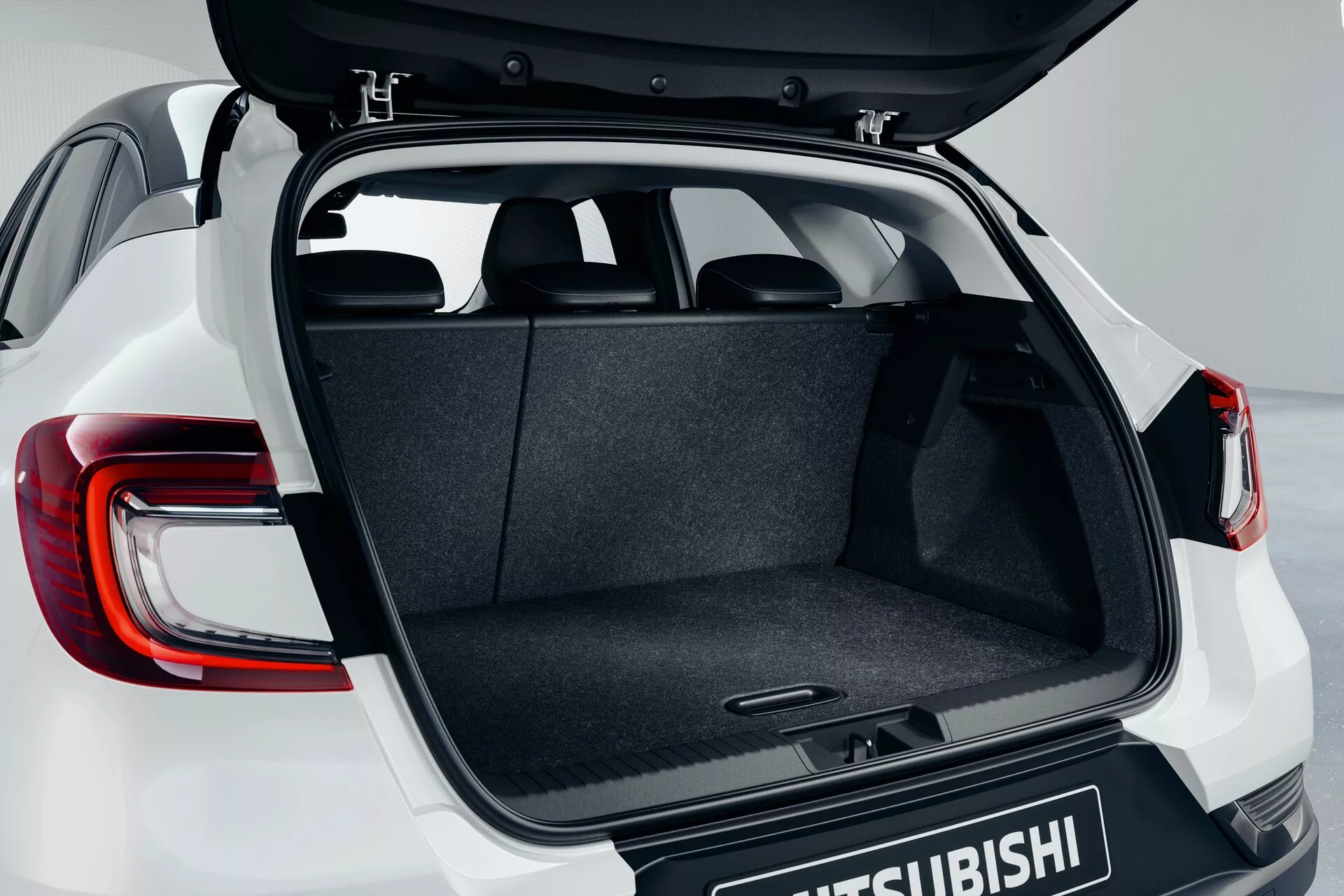 Coffre du Mitsubishi ASX 2023 (2ème génération) montrant le hayon ouvert depuis un angle légèrement surélevé, mettant en avant l'espace de chargement moquetté, le mécanisme de charnière, le plancher plat et les feux arrière LED modernes partiellement visibles avec le logo Mitsubishi sur la porte du coffre.