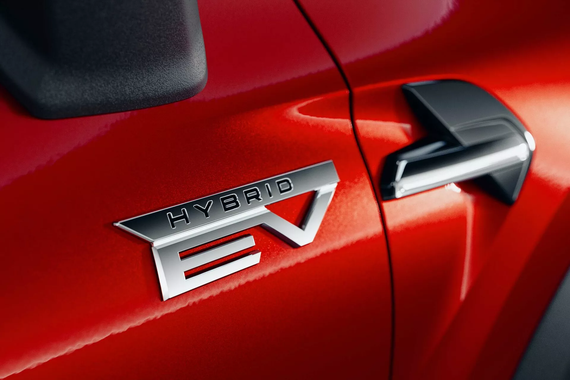 Gros plan détaillé de la Mitsubishi ASX 2023 (2ᵉ génération) en rouge, mettant en avant l'insigne 'HYBRID EV' indiquant la technologie de véhicule hybride électrique. L'insigne 'HYBRID EV' est centré, mettant en valeur la finition métallique et le lettrage précis. Le 'EV' présente un design distinctif avec une flèche traversant le 'V'. Le rétroviseur latéral est partiellement visible dans le coin supérieur droit, suggérant des fonctionnalités technologiques supplémentaires. La finition brillante de la peinture et la couture serrée entre la porte passager avant et l'aile avant suggèrent une qualité de fabrication supérieure.