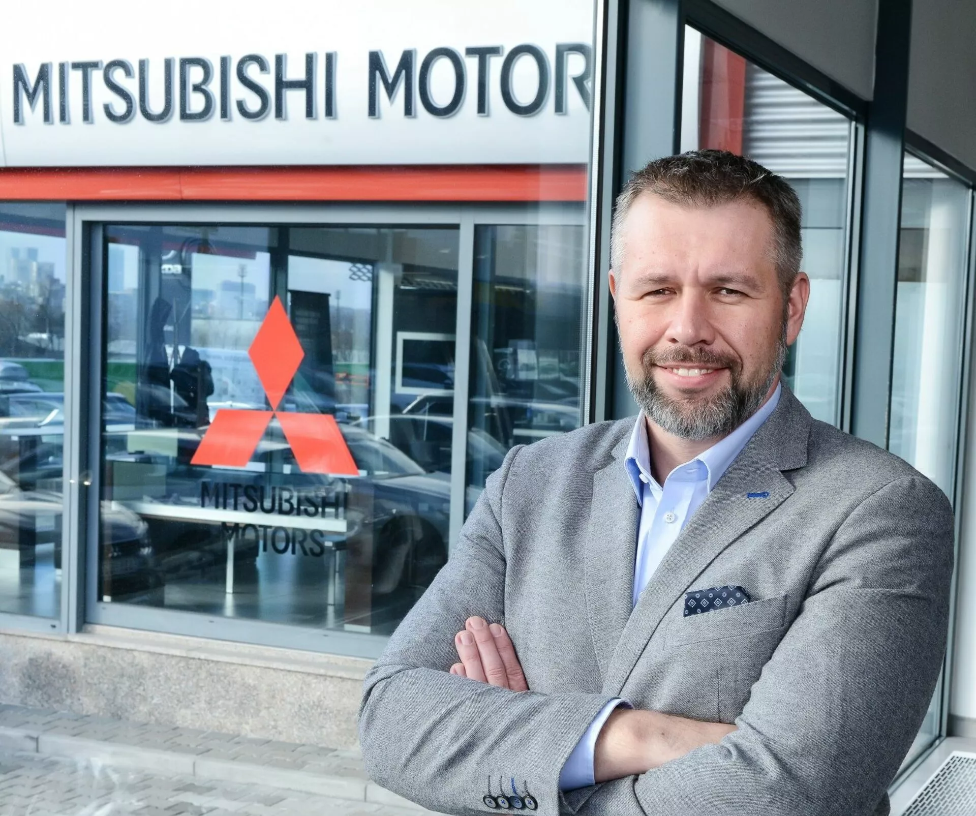 Personne debout les bras croisés devant une concession Mitsubishi Motors, véhicule partiellement visible à travers la fenêtre en verre suggérant un modèle d’exposition en arrière-plan