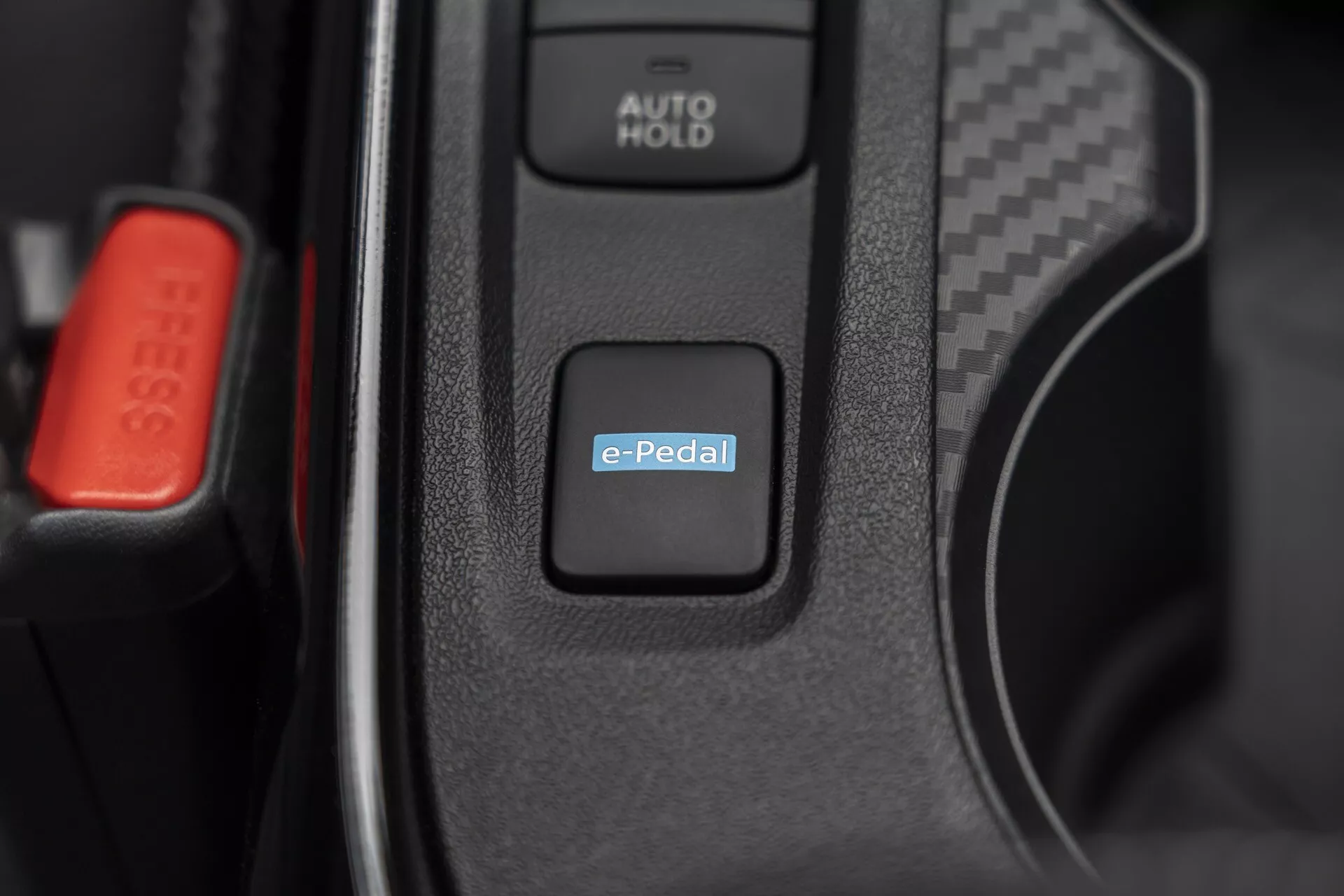 Gros plan sur l'intérieur du Nissan Juke (F16) 2019 mettant en évidence l'interrupteur du système e-Pedal pour le contrôle de la conduite électrique ou hybride, avec le bouton AUTO HOLD et l'attache de ceinture de sécurité rouge visible suggérant une technologie automobile avancée et des caractéristiques intérieures haut de gamme