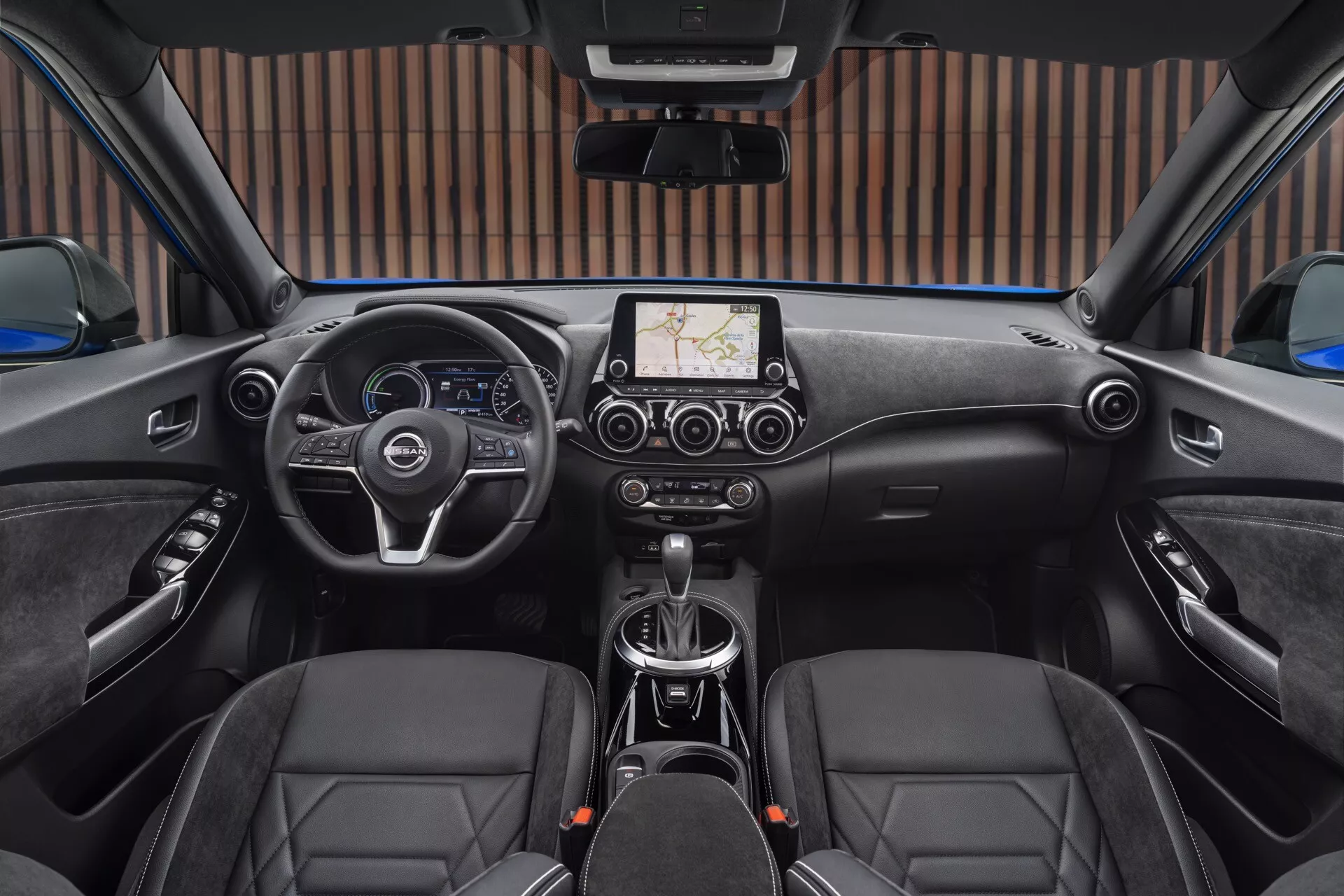 Vue intérieure depuis le siège arrière du Nissan Juke (F16) 2019 montrant le volant à gauche avec des boutons de contrôle, système d'infodivertissement tactile central avec navigation, levier de vitesse manuel, sièges avec coutures en forme de diamant et commandes du toit ouvrant