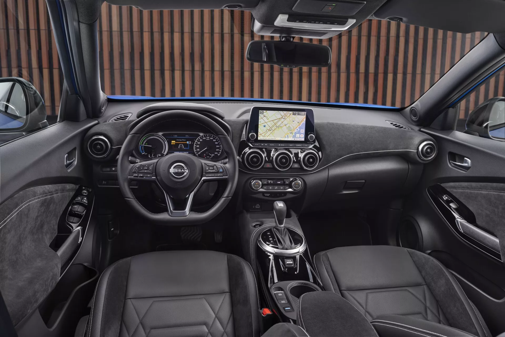 Vue intérieure du Nissan Juke F16 de 2019 depuis la perspective du conducteur, montrant un volant moderne à trois branches avec des boutons de commande, un tableau de bord combiné numérique et analogique, un système d'infodivertissement central avec écran tactile et navigation, contrôle climatique bi-zone, transmission automatique, sièges baquets en cuir et frein de stationnement électronique.