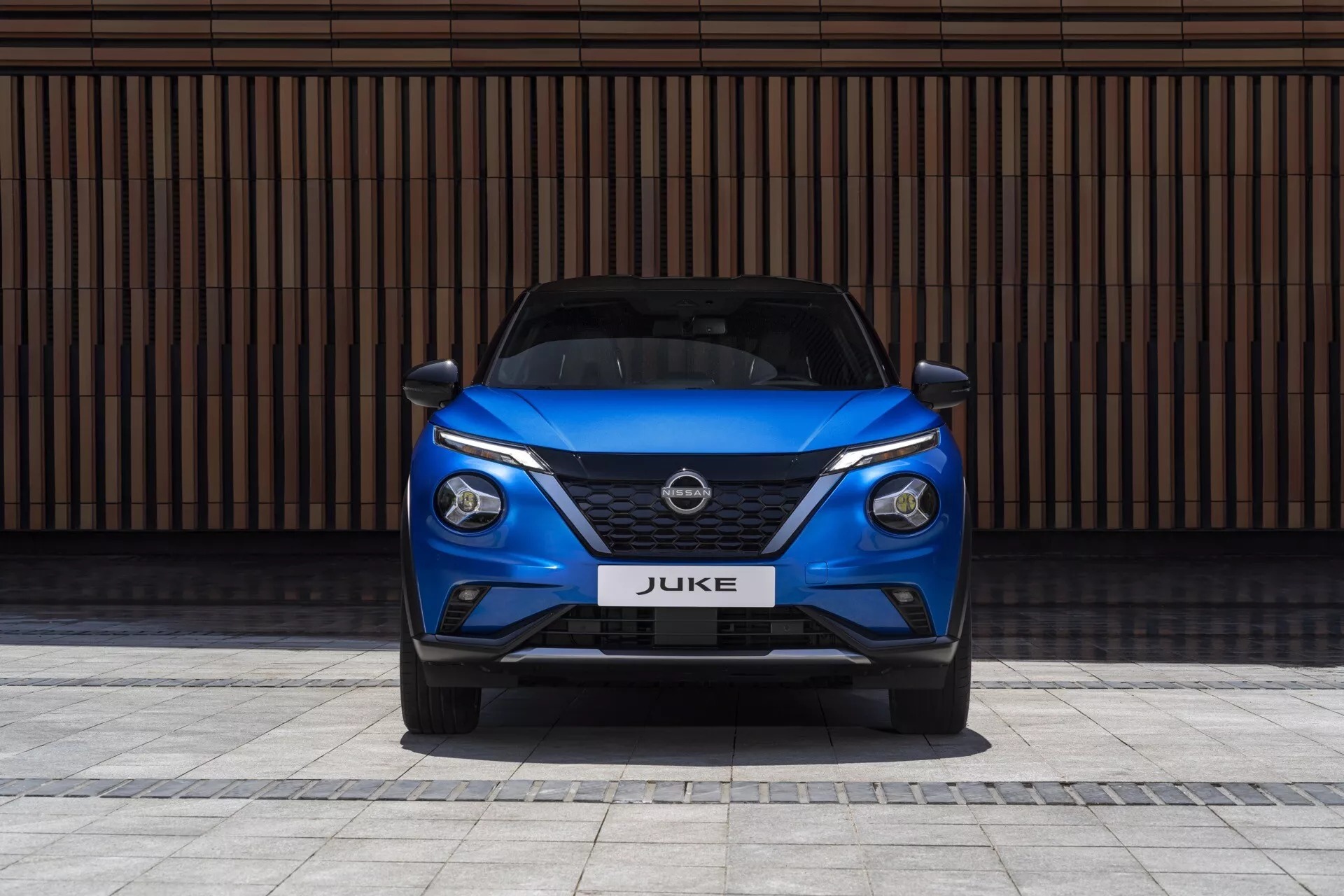 Vue de face d'un Nissan Juke F16 de 2019 en bleu, présentant des phares LED élégants, une calandre V-motion, et un pare-chocs sportif sur un fond de mur à lignes horizontales