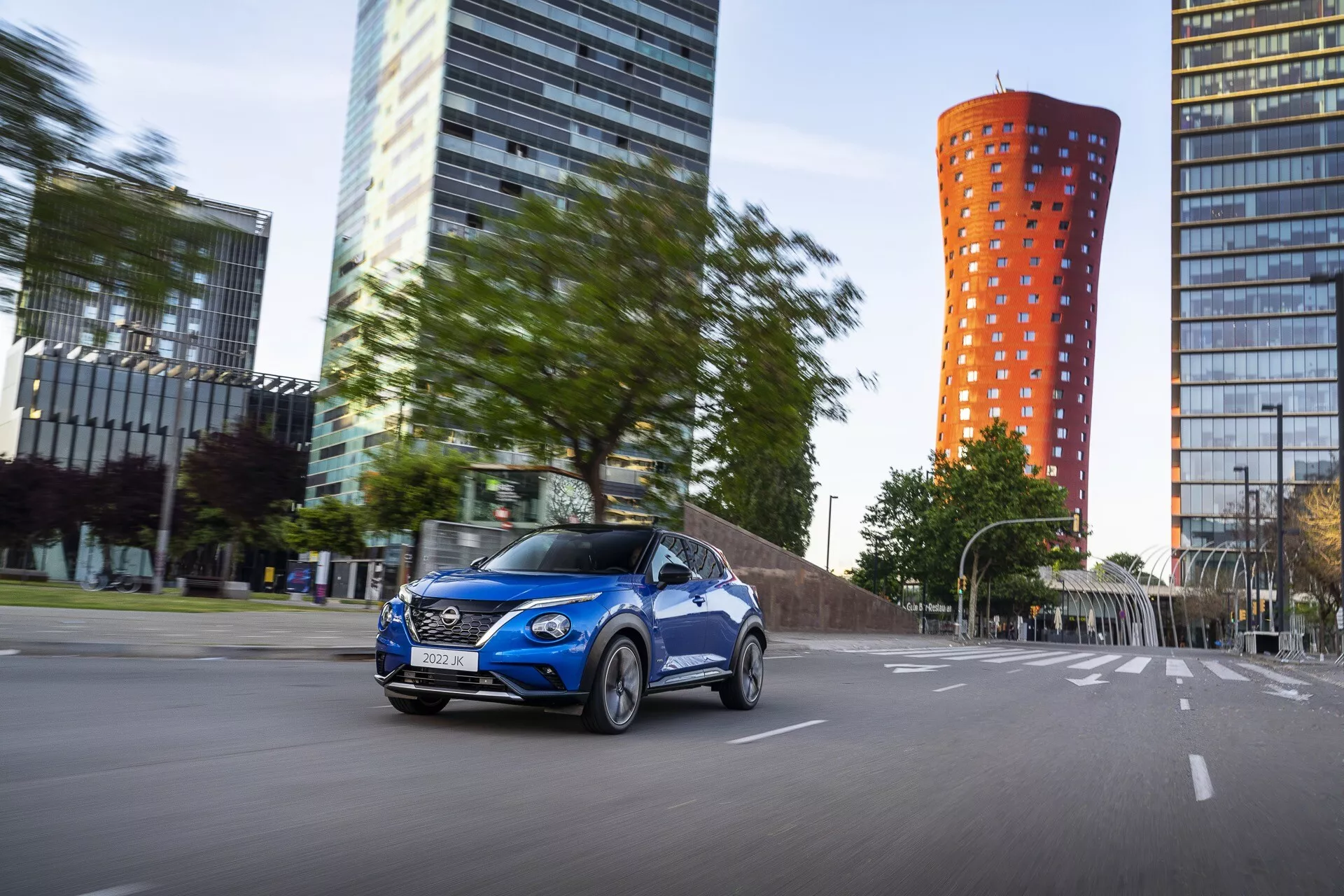 Nissan Juke F16 2019 en bleu vibrant, en mouvement sur route urbaine avec vue dynamique avant trois quarts côté conducteur, mettant en avant jantes alliage, carrosserie profilée et phares activés sur fond de ville moderne avec effet de flou de mouvement.
