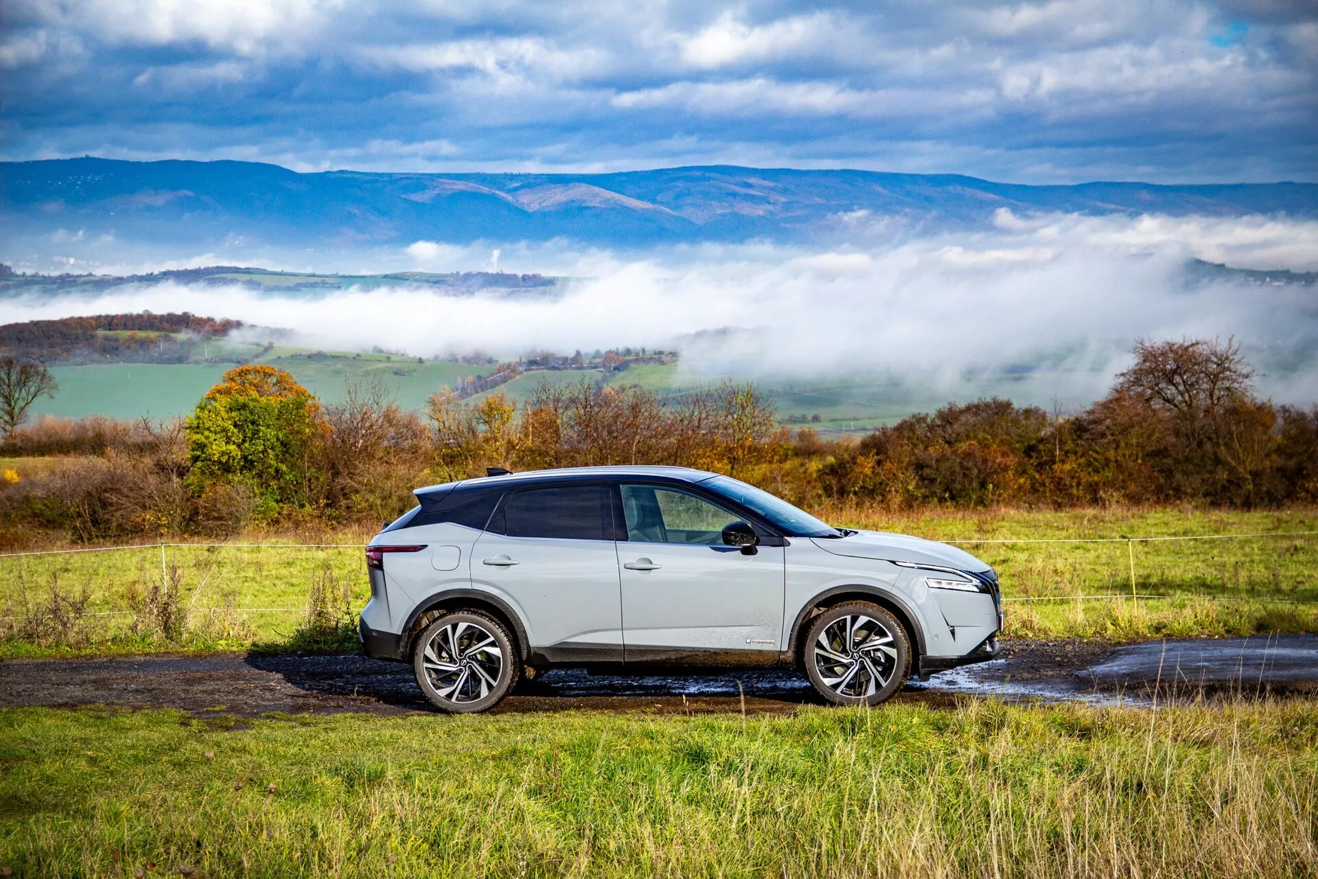 Vue de profil latéral du Nissan Qashqai 2021, un SUV moderne avec une finition argentée ou gris clair brillante, de grandes roues en alliage multi-rayons, des vitres teintées et une ligne de toit inclinée, garé sur une route de campagne mouillée avec des flaques d'eau. Le design élégant de la voiture, les unités d'éclairage modernes et les clignotants intégrés dans les rétroviseurs latéraux sont clairement visibles sur un fond de collines ondulées et de nuages bas.