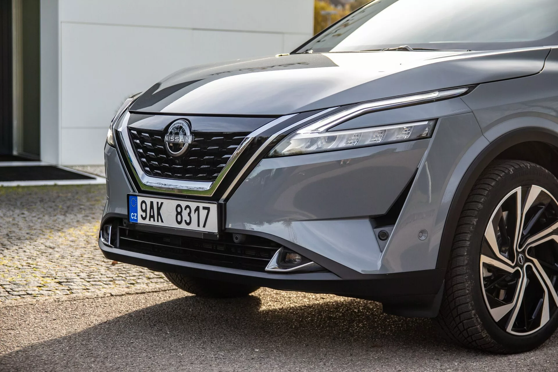 Gros plan sur le côté avant gauche d'une voiture moderne, présentant des phares LED élégants, une grille proéminente avec des accents chromés, un capot sculpté, un garde-boue bombé, des jantes en alliage à plusieurs branches et un rétroviseur conducteur avec clignotants intégrés, reflétant le design dynamique et les capacités de maniabilité potentielles de la voiture.