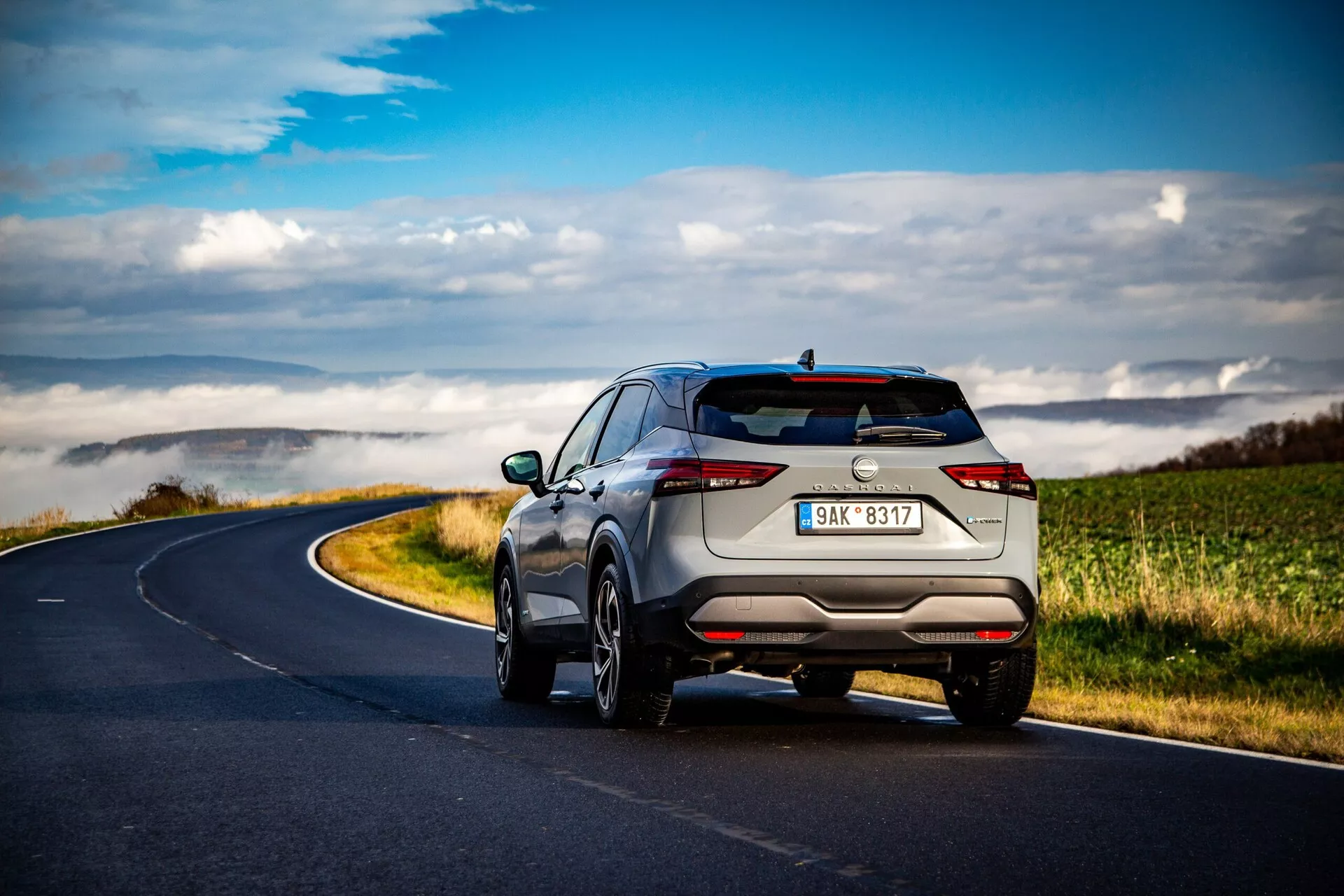 SUV Nissan Qashqai 2021 pris sous un angle arrière trois-quarts sur une route rurale sinueuse sous des nuages épars, mettant en valeur un design élégant et moderne avec des feux arrière fins et horizontaux, des tuyaux d'échappement discrets, des roues en alliage à plusieurs rayons, des rails de toit et des capteurs de stationnement avancés. La couleur métallique claire souligne son aspect haut de gamme.