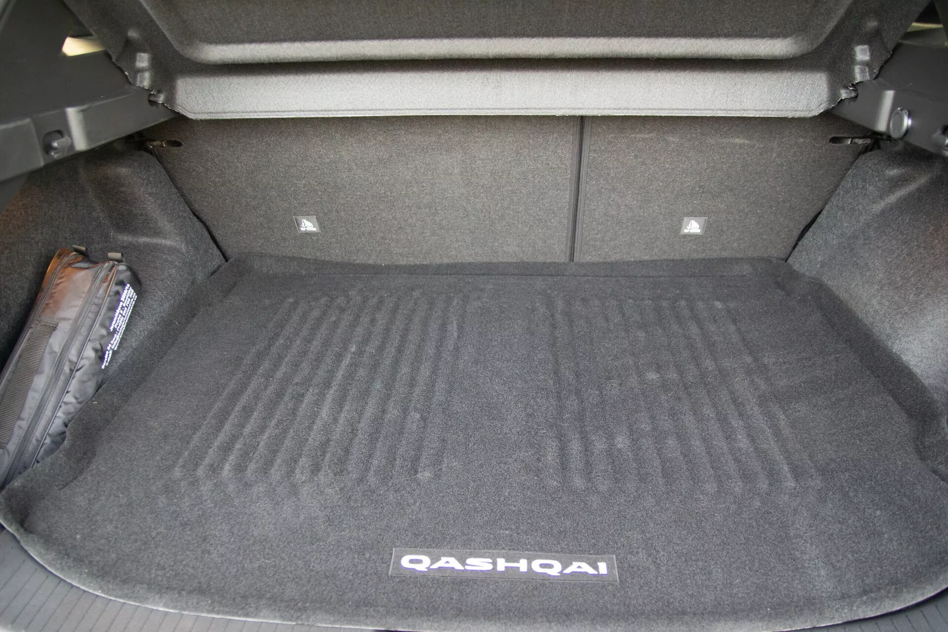 Image montrant l'espace de coffre ouvert d'une Nissan Qashqai 2021 depuis un angle arrière. Le coffre, dépourvu de toute cargaison, révèle sa surface recouverte de moquette, ses parois latérales et les sièges arrière en position verticale. Une zone légèrement surélevée à l'avant du coffre suggère des composants mécaniques sous-jacents ou un support pour les sièges arrière. Une petite cloison de rangement est visible à l'arrière du coffre. À gauche, il y a un article d'équipement d'urgence, probablement un cric de pneu ou une trousse de premiers secours. L'image met en évidence la capacité spacieuse et la conception fonctionnelle du coffre du véhicule.