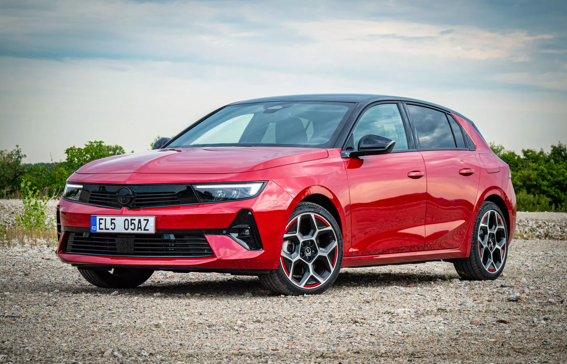 Hatchback rouge Opel Astra 2022 photographié depuis une vue frontale trois quarts montrant les phares LED, la grille avant proéminente, les roues en alliage à multiples branches et le profil latéral élégant sur une surface de gravier avec une lumière solaire diffuse mettant en lumière les caractéristiques modernes de la voiture