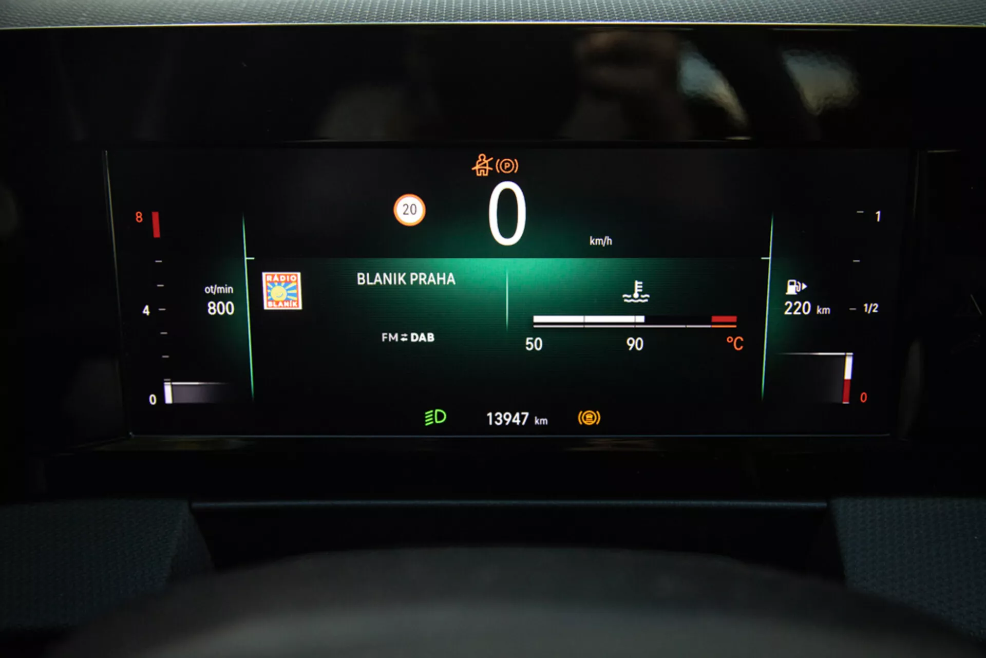 Vue du tableau de bord numérique de l'Opel Astra 2022 mettant en évidence le bloc d'instruments avec le régime moteur, le niveau de batterie, le compteur de vitesse, la jauge de carburant, la température extérieure, la station de radio 'BLANIK PRAHA', et la lecture de l'odomètre depuis la perspective du siège du conducteur.