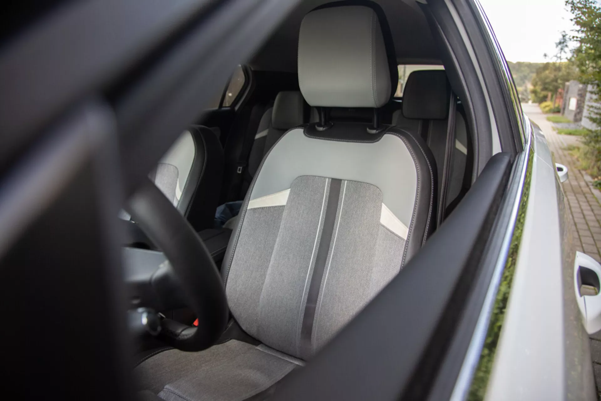 Vue rapprochée de l’intérieur côté conducteur de l’Opel Astra 2022 depuis un angle latéral extérieur montrant le siège conducteur en tissu bicolore, appuie-tête intégré, partie du volant avec commandes, aperçu du tableau de bord, ceinture de sécurité attachée, intérieur de la porte avec contrôles électriques des vitres potentiels, et boîtier de rétroviseur latéral de couleur carrosserie mettant en avant l’ergonomie, la sécurité et les fonctionnalités de commodité.