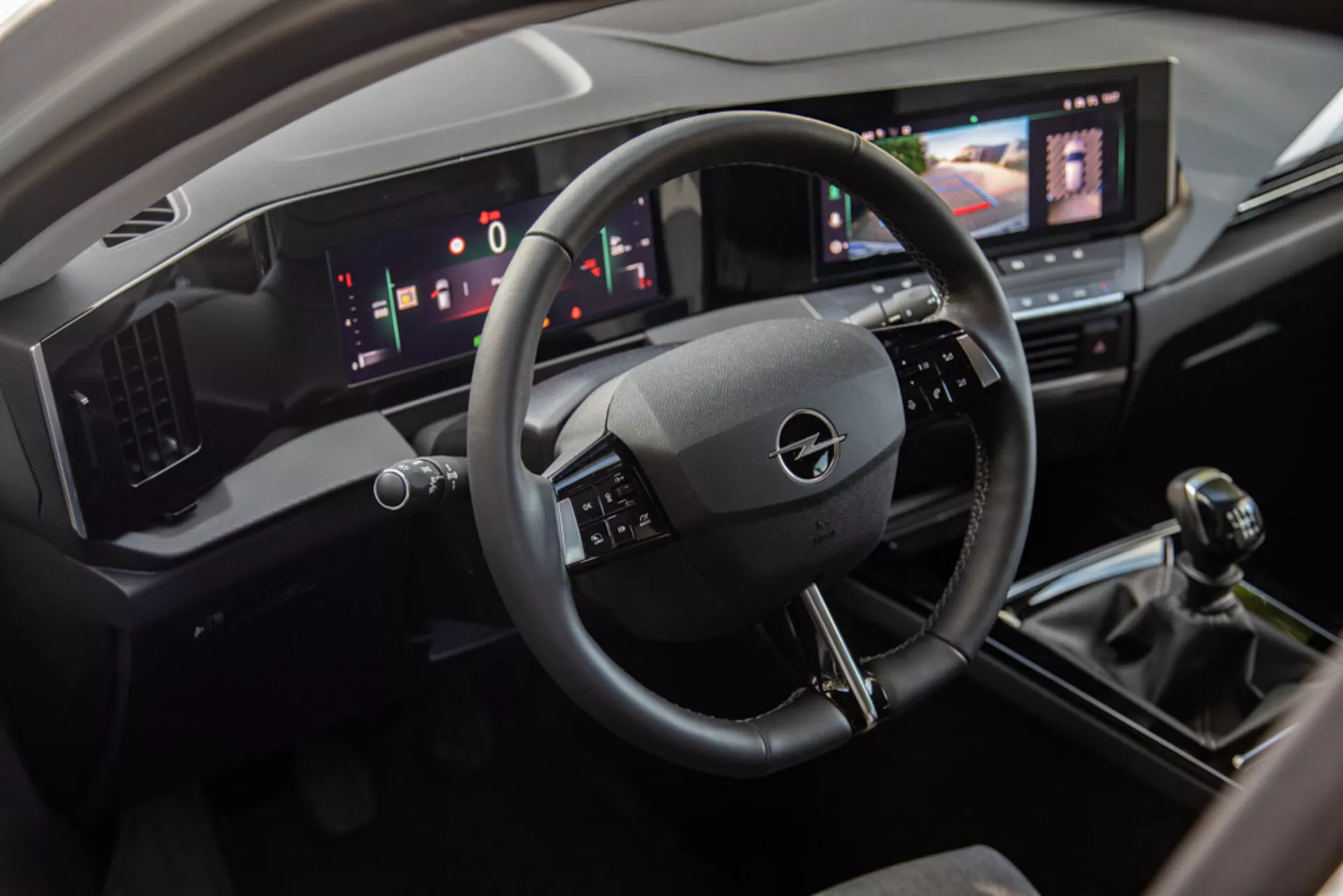 Vue intérieure de l'Opel Astra 2022 montrant la zone du conducteur avec un volant à trois branches, un tableau de bord numérique et une console centrale incluant un système d'infodivertissement tactile, un levier de vitesse manuel et des commandes de climatisation modernes