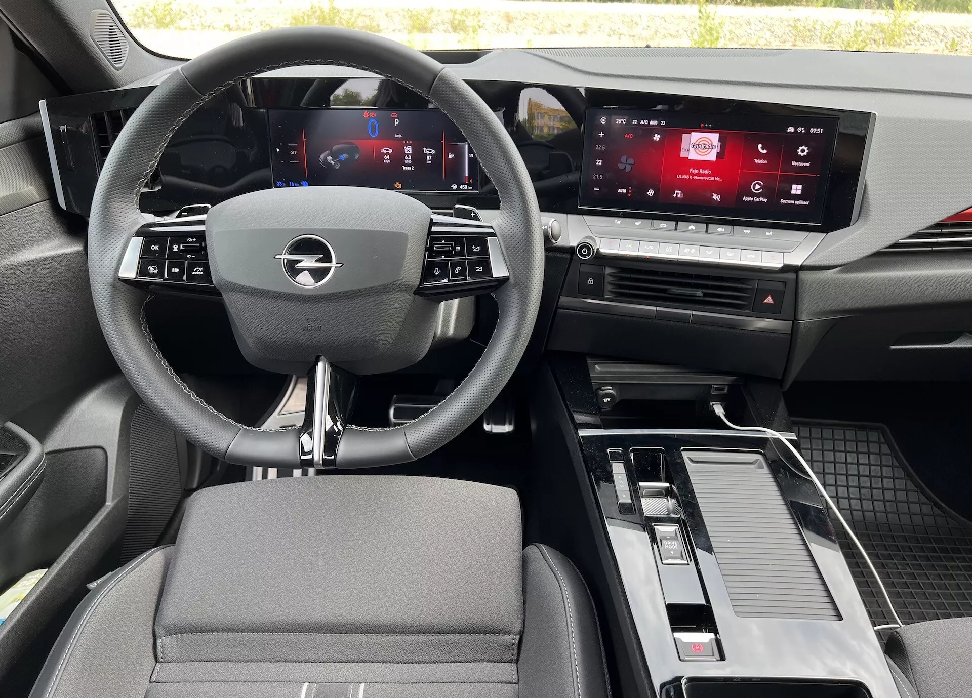 Vue intérieure de l'Opel Astra 2022 depuis le siège arrière, montrant le volant à trois branches avec commandes intégrées, tableau de bord numérique, écran tactile de l'infotainment, console centrale avec levier de vitesse et rangement, et un mélange de matériaux en tissu et effet métal