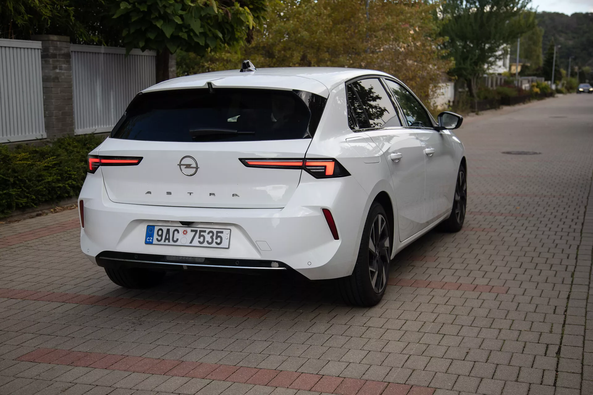 Sedan moderne Opel Astra 2022 en blanc, garée sur une surface pavée de briques entrelacées, affichant un angle arrière de trois quarts avec un accent sur les feux arrière LED haute technologie, la caméra de recul, les capteurs de stationnement, profil latéral partiel incluant la roue et la porte arrière droite, reliés par une barre lumineuse horizontale à travers le couvercle du coffre, équipée d'une antenne aileron de requin pour une connectivité améliorée, adaptée pour illustrer le design arrière du véhicule et la technologie automobile moderne
