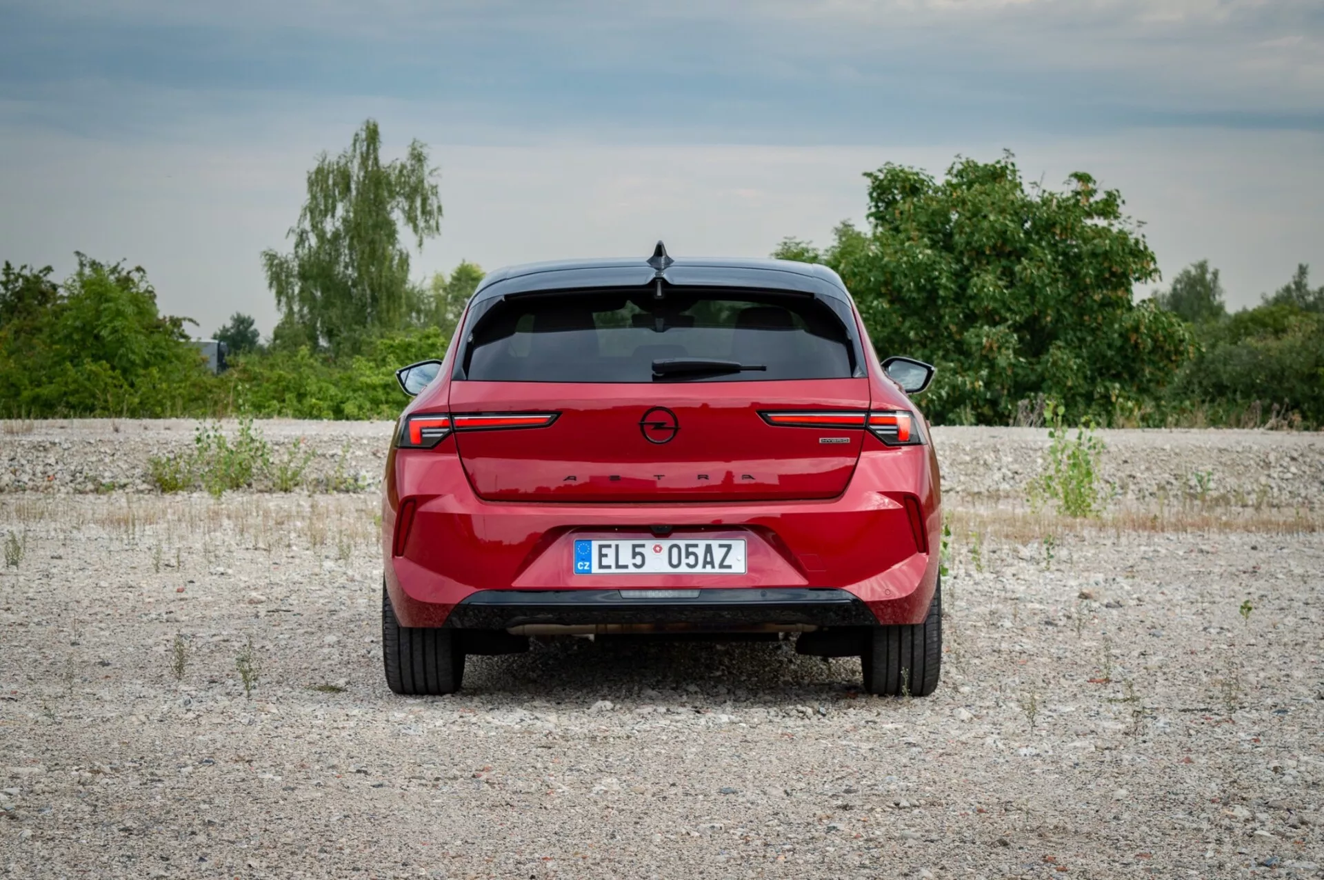 Opel Astra 2022 photographiée de l'arrière sur une surface de gravier montrant les feux arrière avec une barre lumineuse de connexion, des capteurs de stationnement et l'absence de ports d'échappement indiquant un potentiel modèle électrique ou hybride, sur un fond de végétation et de ciel légèrement nuageux.