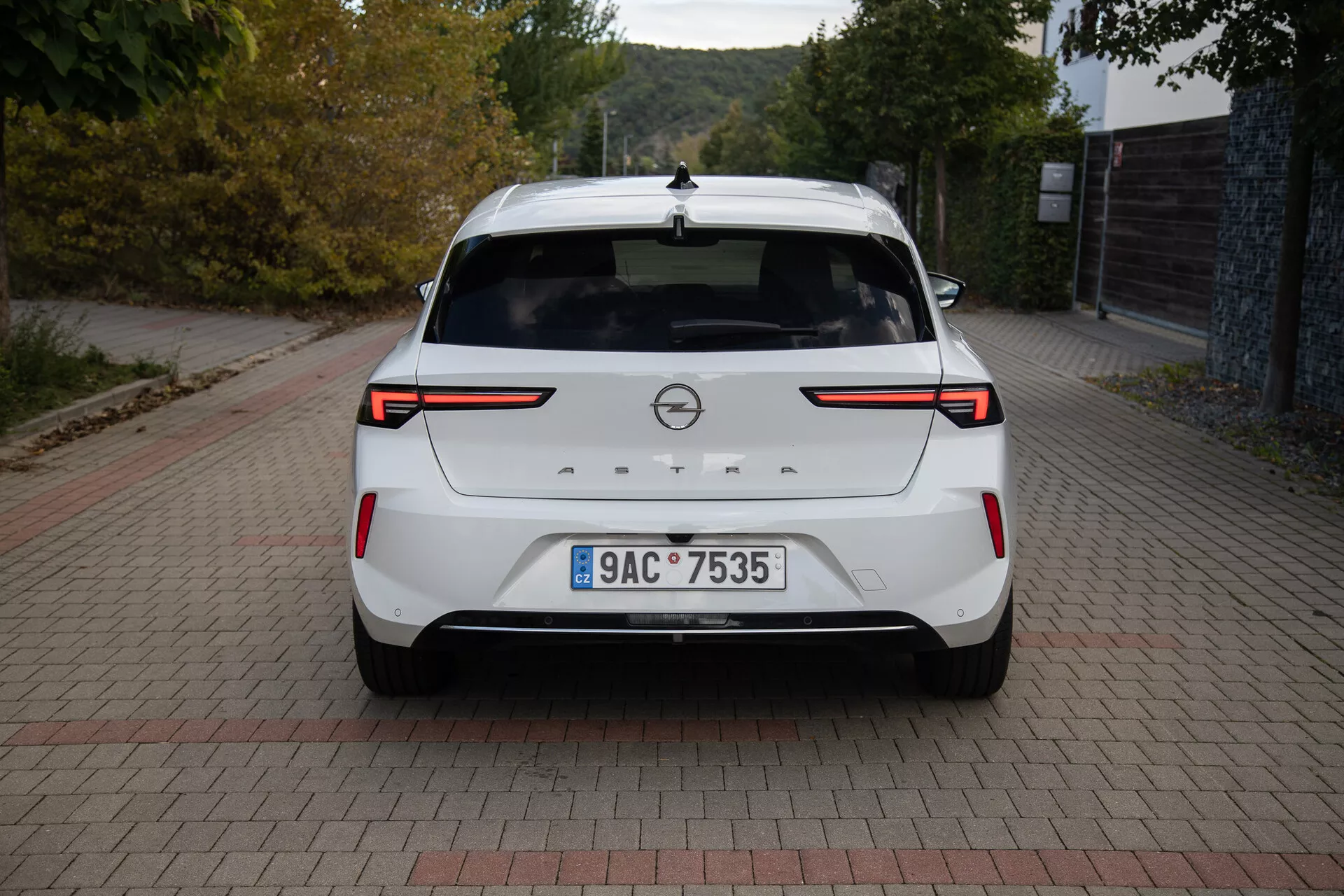 Vue arrière d'une Opel Astra 2022 blanche montrant des feux arrière LED, des capteurs/caméra de parking arrière, un badge de marque, une plaque d'immatriculation, un aileron arrière, un feu stop surélevé, une antenne aileron de requin et une finition de peinture propre, stationnée sur une surface à motif de briques à la lumière du jour.