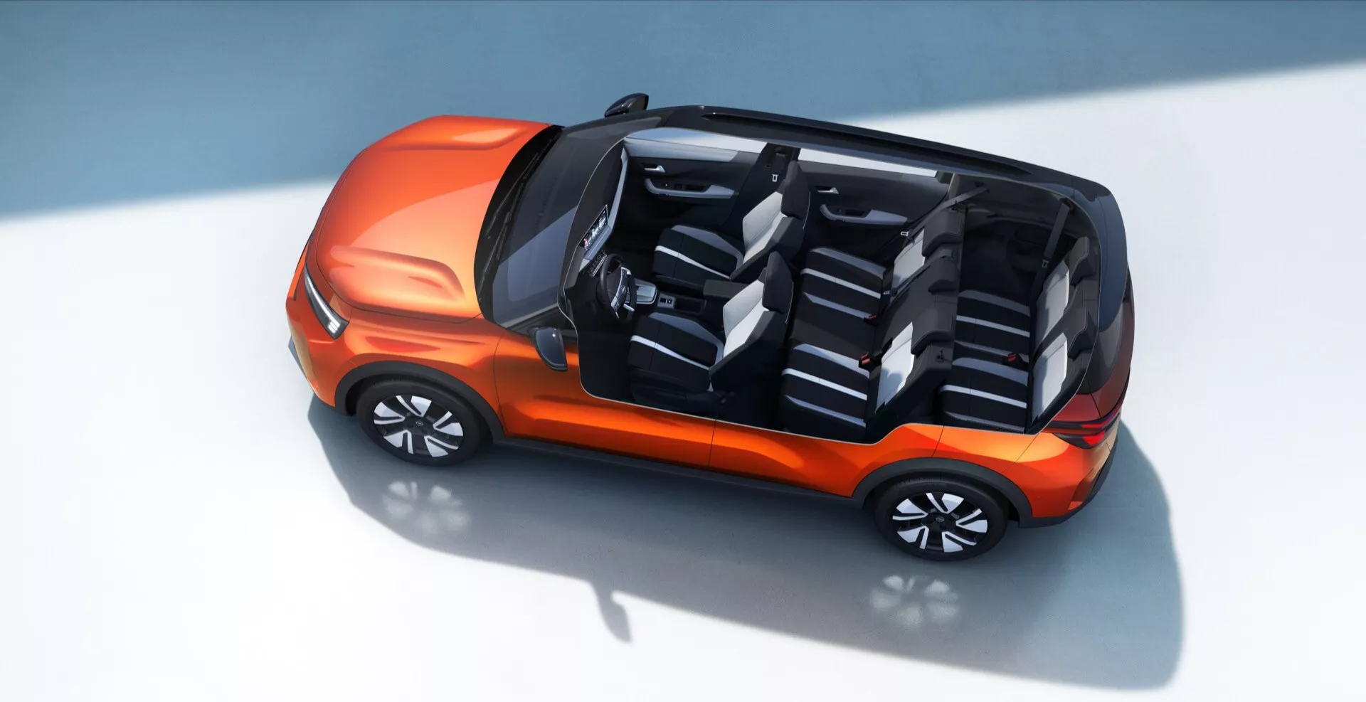 Vue de haut et légèrement de face de l'Opel Frontera 2024 (3ème génération), mettant en valeur son extérieur orange vif et son agencement intérieur luxueux. La voiture dispose d'une disposition de sièges à deux rangées avec quatre sièges ergonomiques en dégradé de gris, soulignant le confort des passagers. L'absence de levier de vitesses traditionnel suggère des technologies avancées telles que le drive-by-wire ou des capacités autonomes. Le fond propre et neutre et les détails des ombres mettent en valeur le design moderne et la posture dynamique du véhicule.